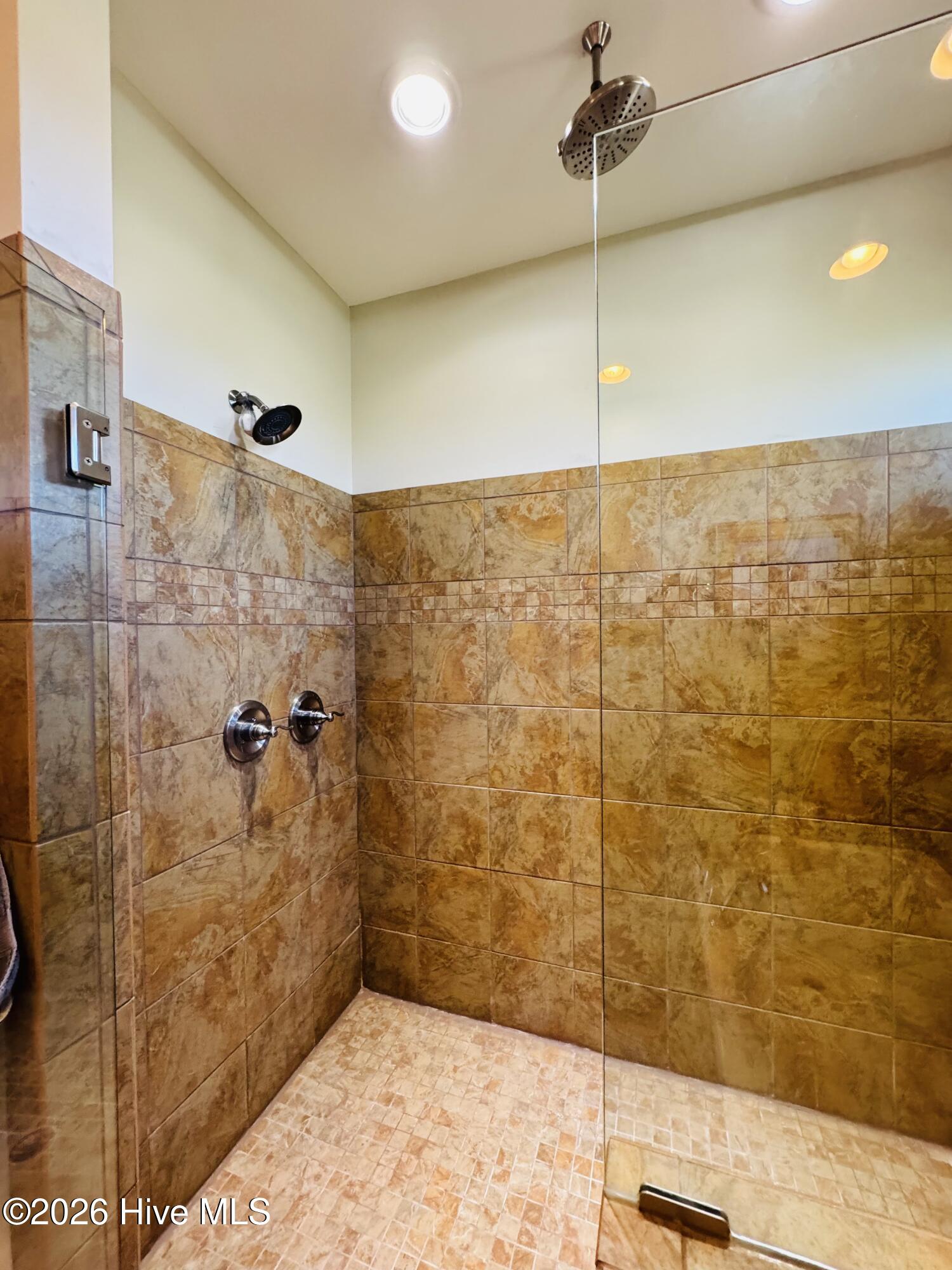 Walk-in shower with warm stone-look tile, glass enclosure, and rainfall and wall-mounted showerheads.