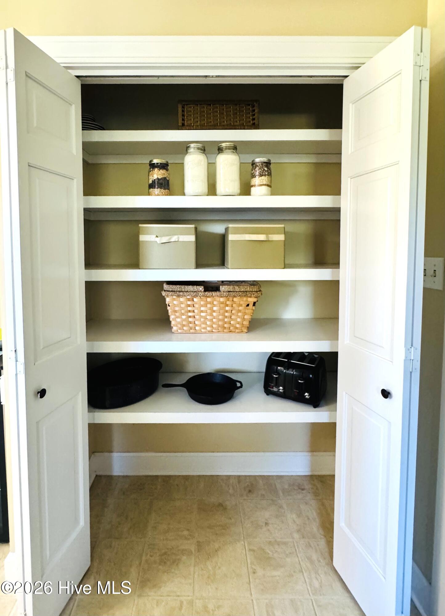 Open pantry closet with white doors, neatly arranged shelves, and beige tile flooring.