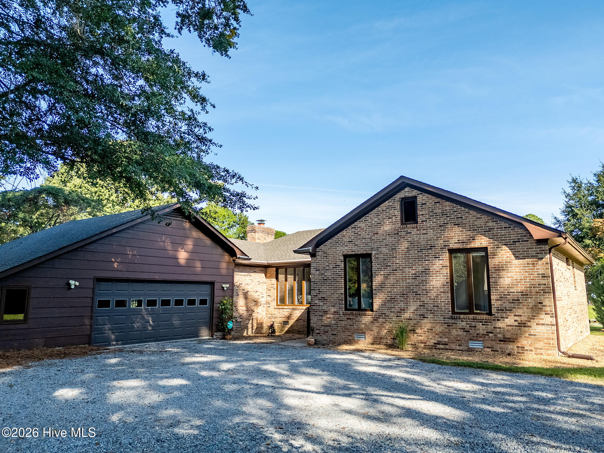 Brick house with dark attached garage and a gravel driveway beneath leafy trees.