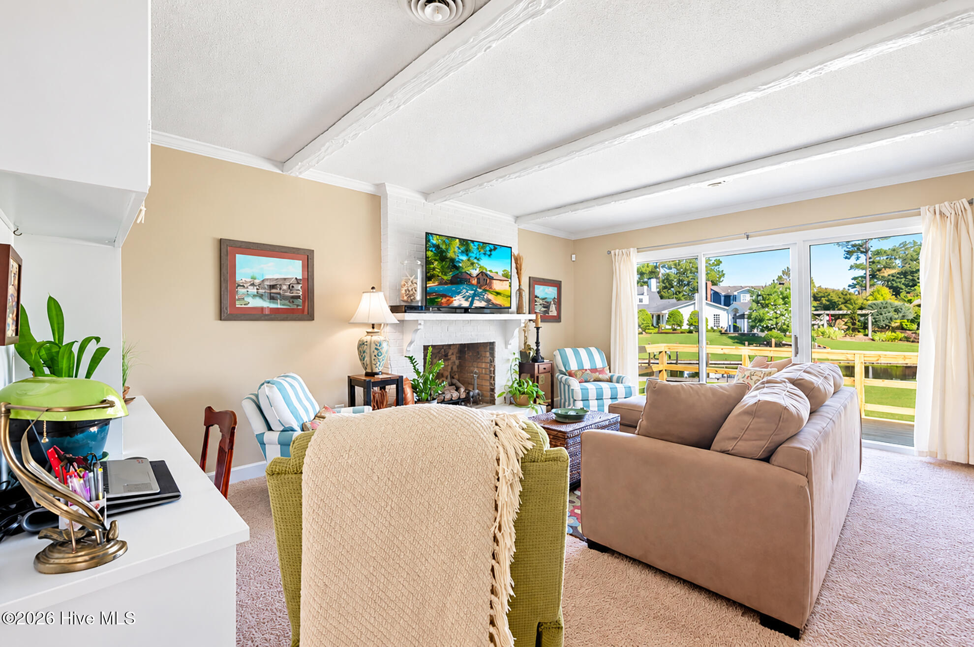 Living room with a brick fireplace, white-beamed ceiling, and sliding doors overlooking a deck and water.