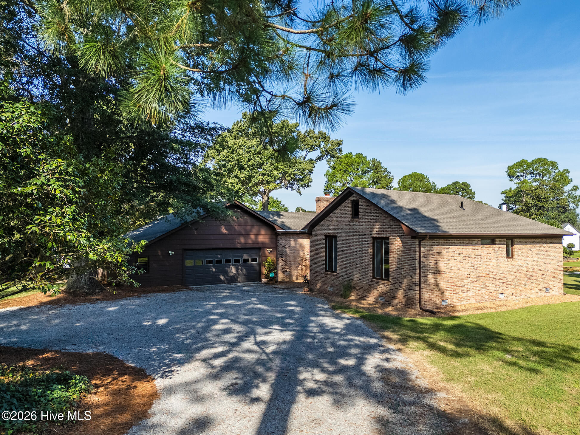 Brick home with an attached garage and broad gravel driveway beneath mature trees.