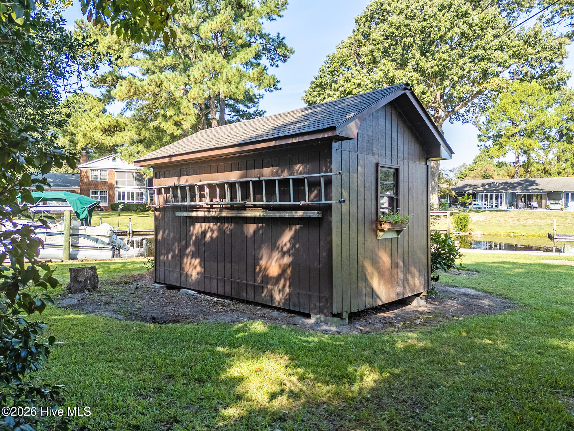 Dark brown backyard shed beside a canal, with a dock and covered boat in the background.
