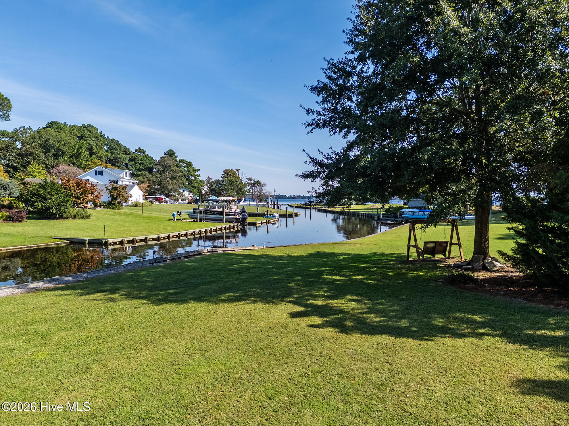 Waterfront lawn beside a canal, with docks, boats, and a swing beneath a large tree.