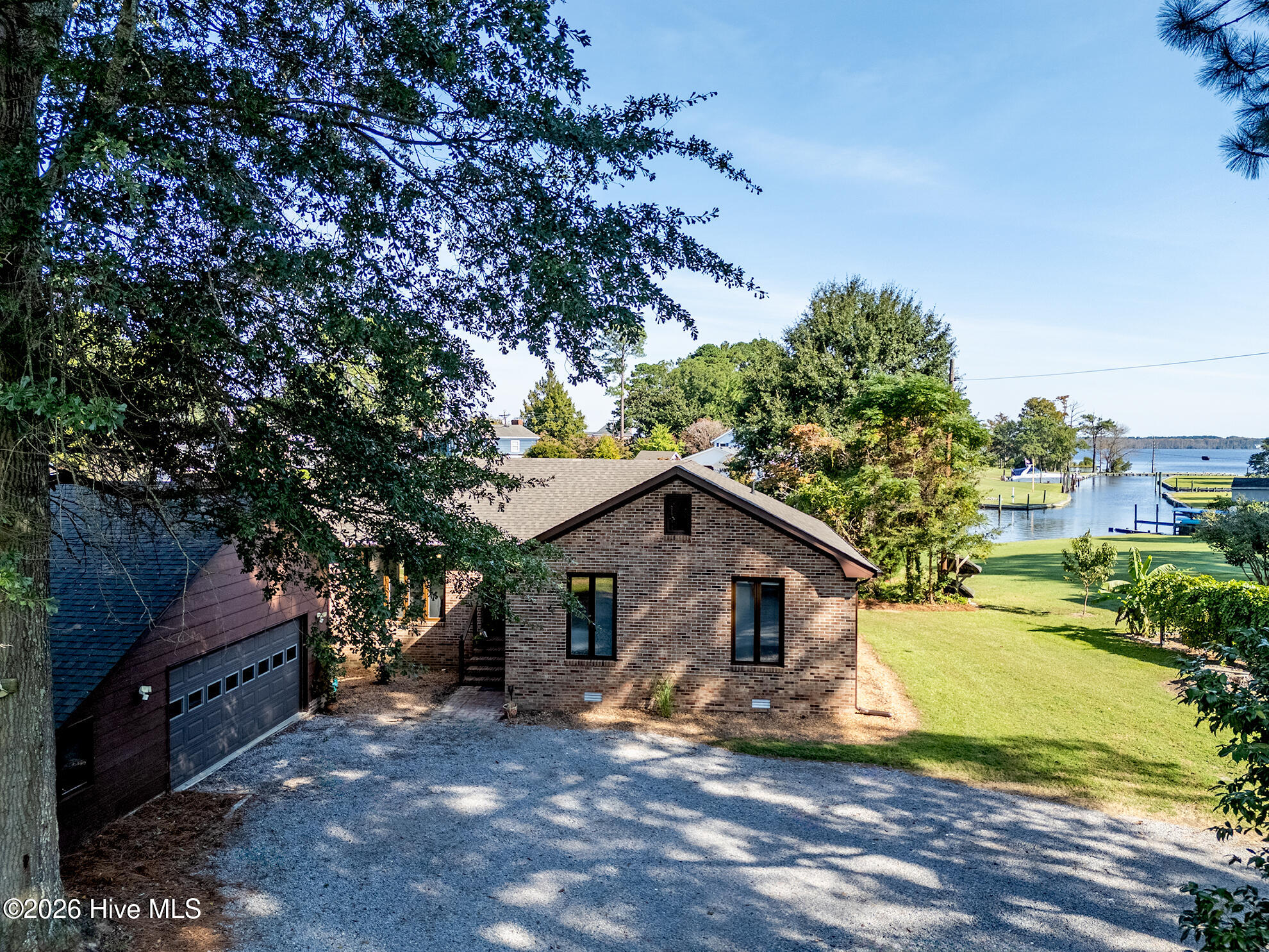 Brick waterfront house with attached garage, broad gravel driveway, green lawn, and dock.