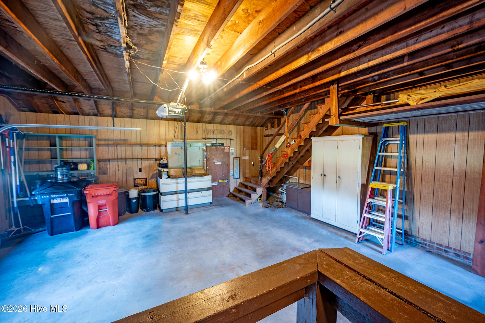 Wood-paneled garage with exposed rafters, a staircase to a loft, storage bins, and a ladder.