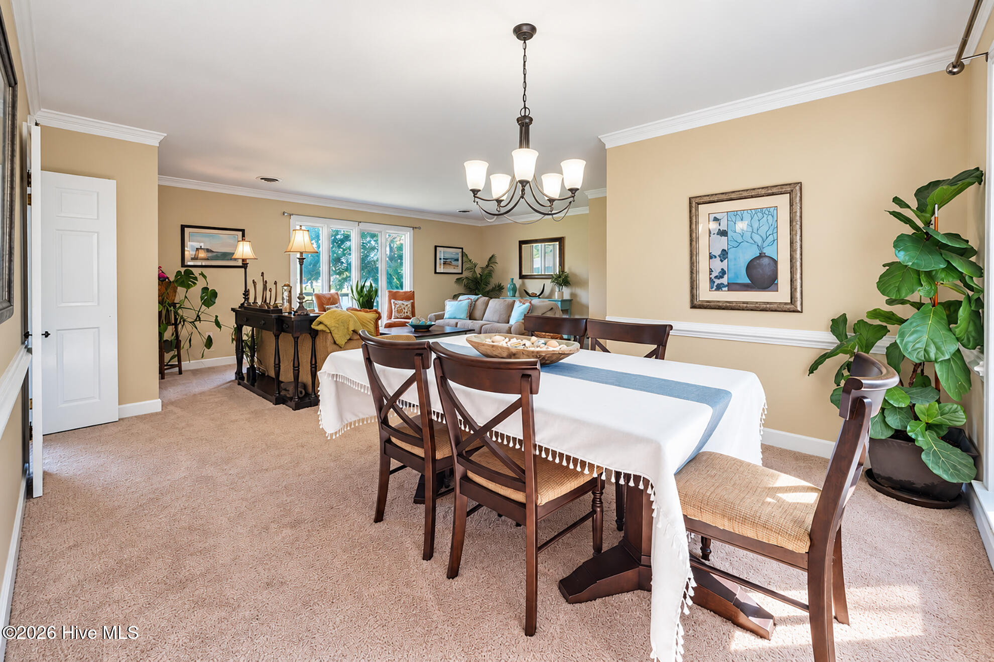 Dining room with a dark wood table, chandelier, and leafy plants beside a bright living area.