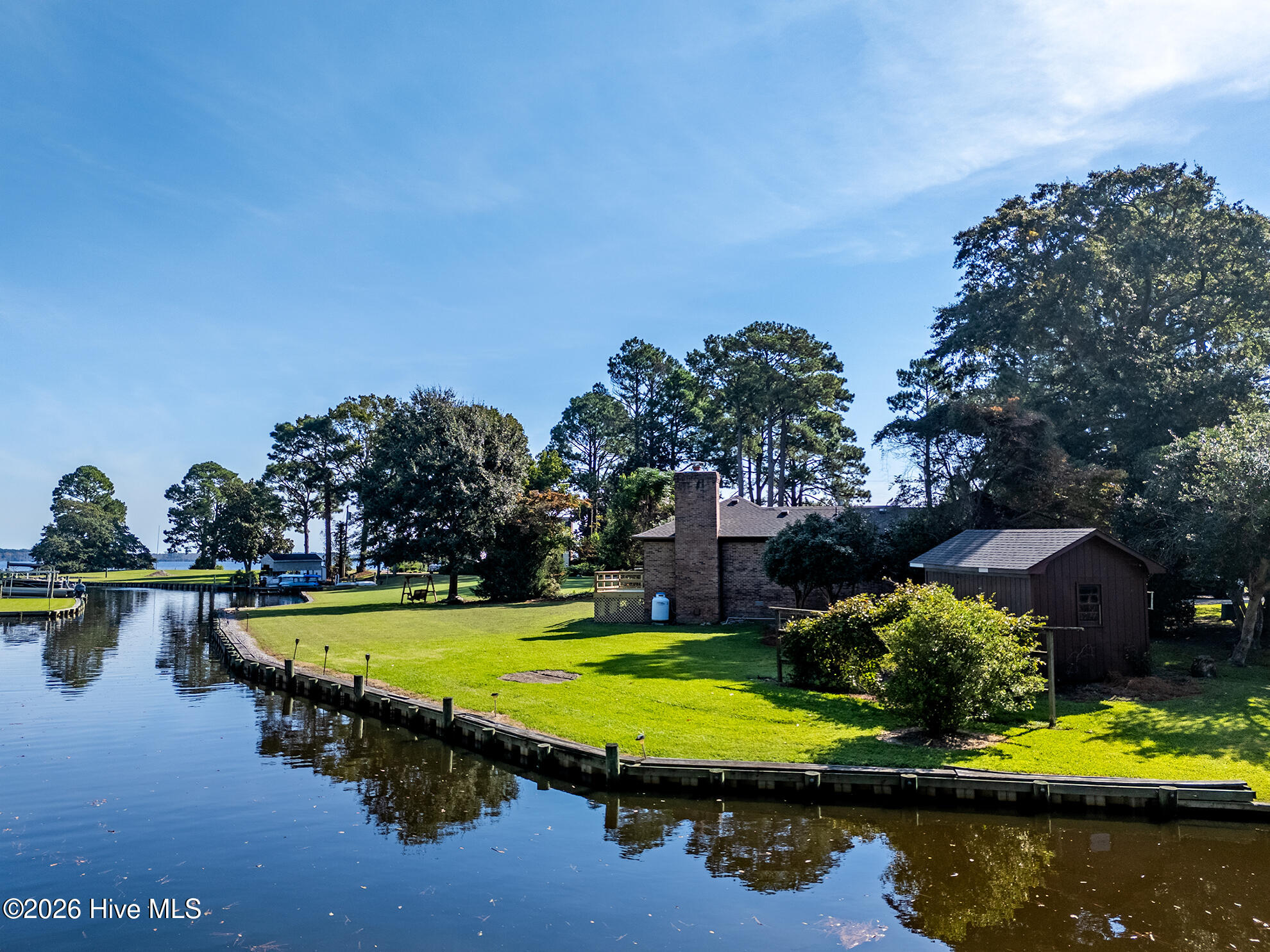 Brick waterfront home beside a calm canal, with a broad lawn, curved bulkhead, and detached shed.