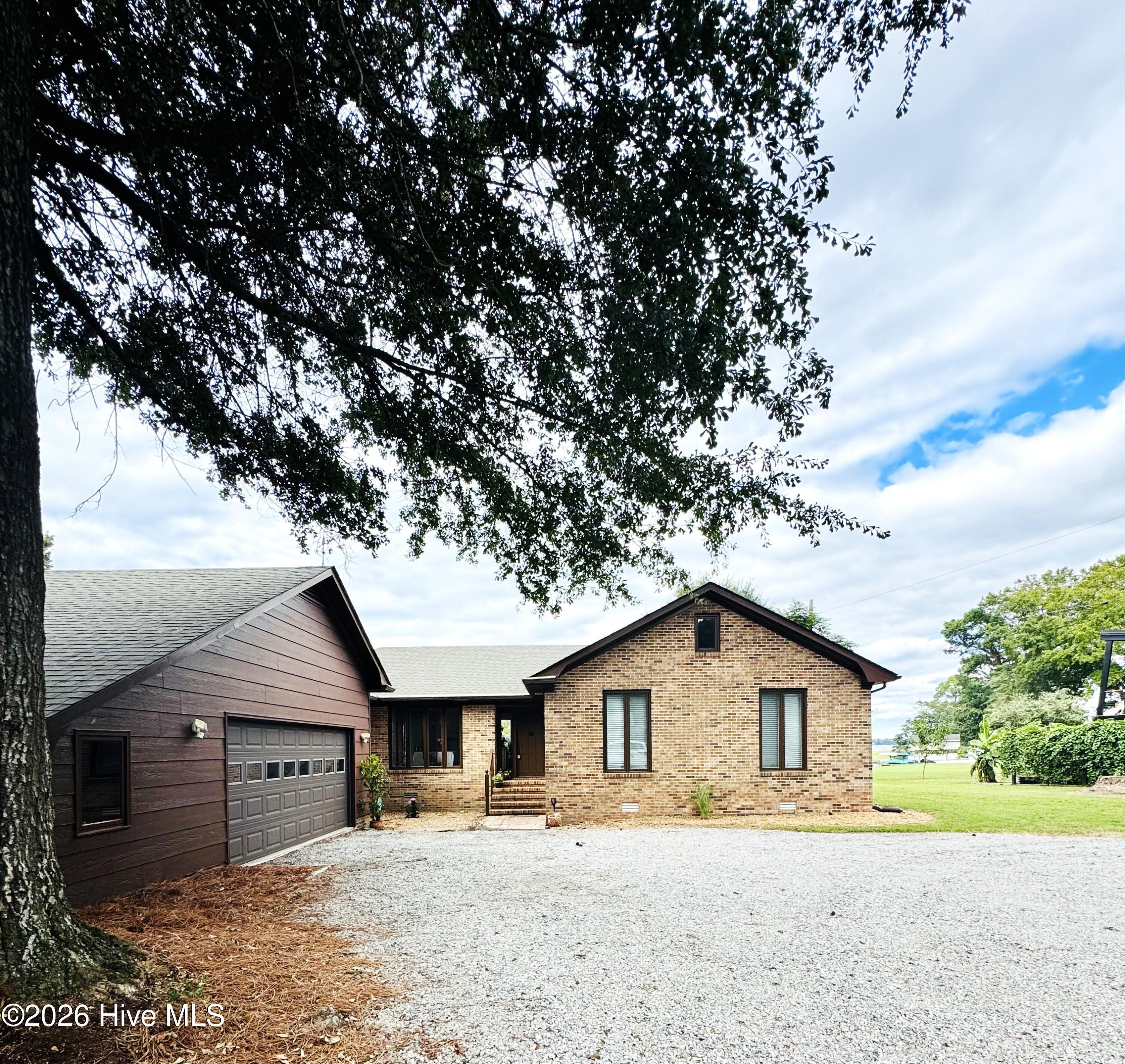 Brick house with attached garage, broad gravel driveway, and a large shade tree in front.