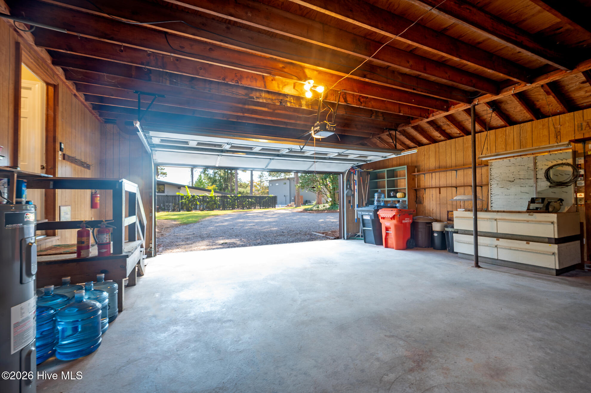 Garage with exposed wood beams, concrete floor, and open door to a gravel driveway.