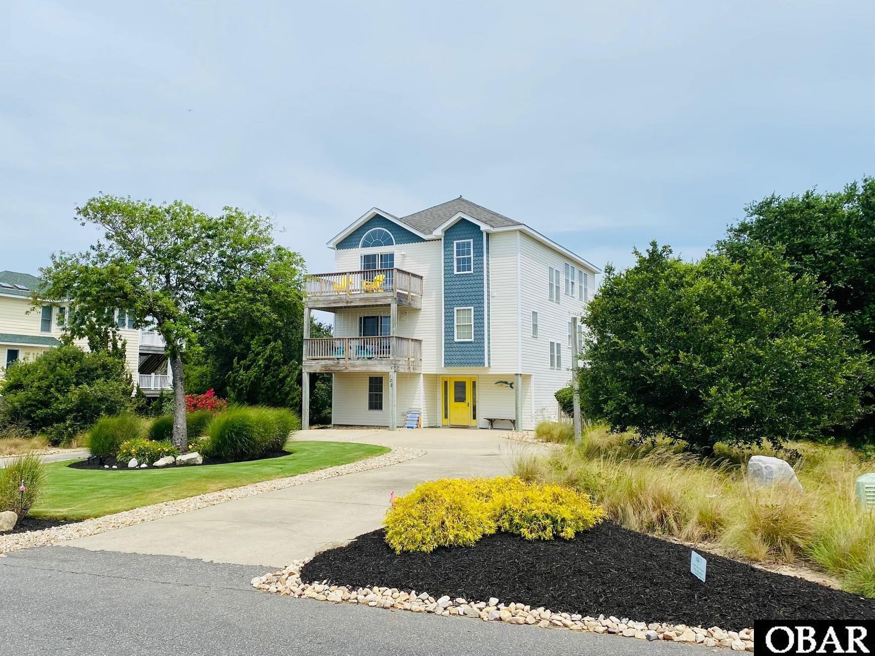 Three-story coastal-style house with blue accents, a yellow front door, and landscaped yard.