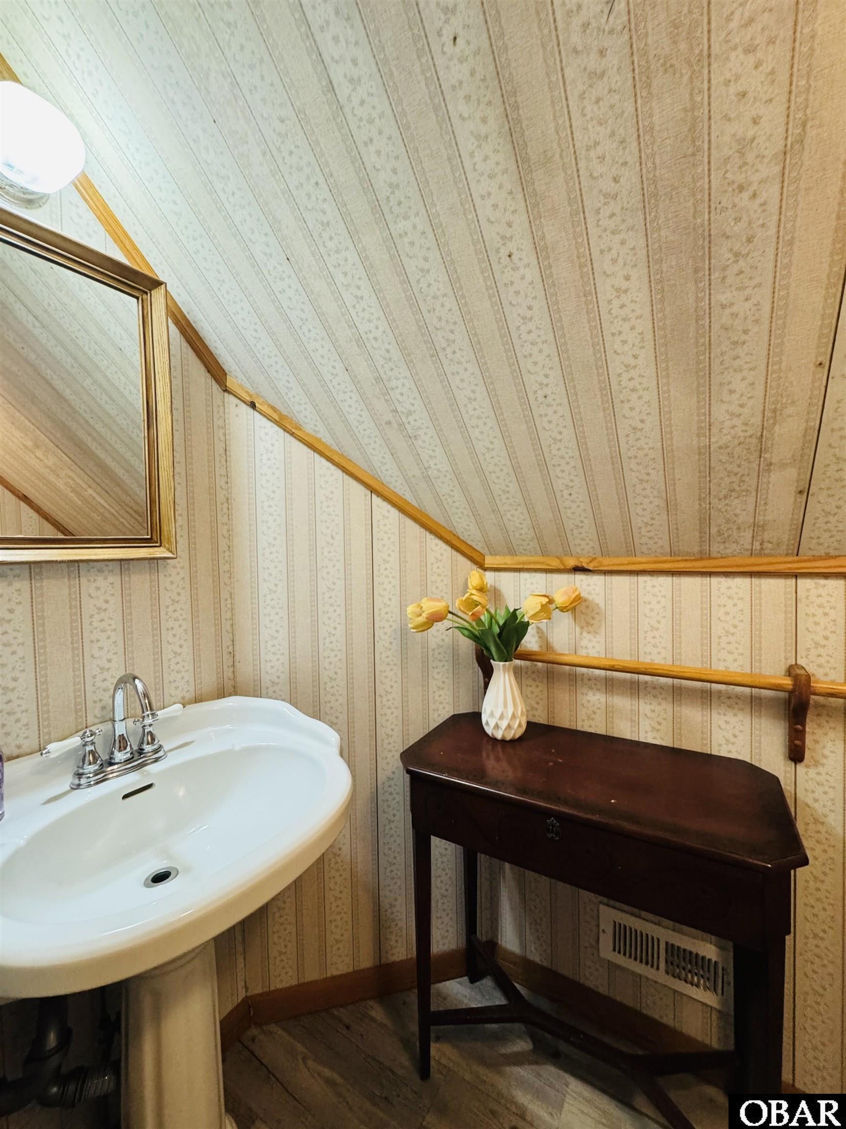 Bathroom with a pedestal sink, framed mirror, sloped ceiling, and console table topped with yellow tulips.