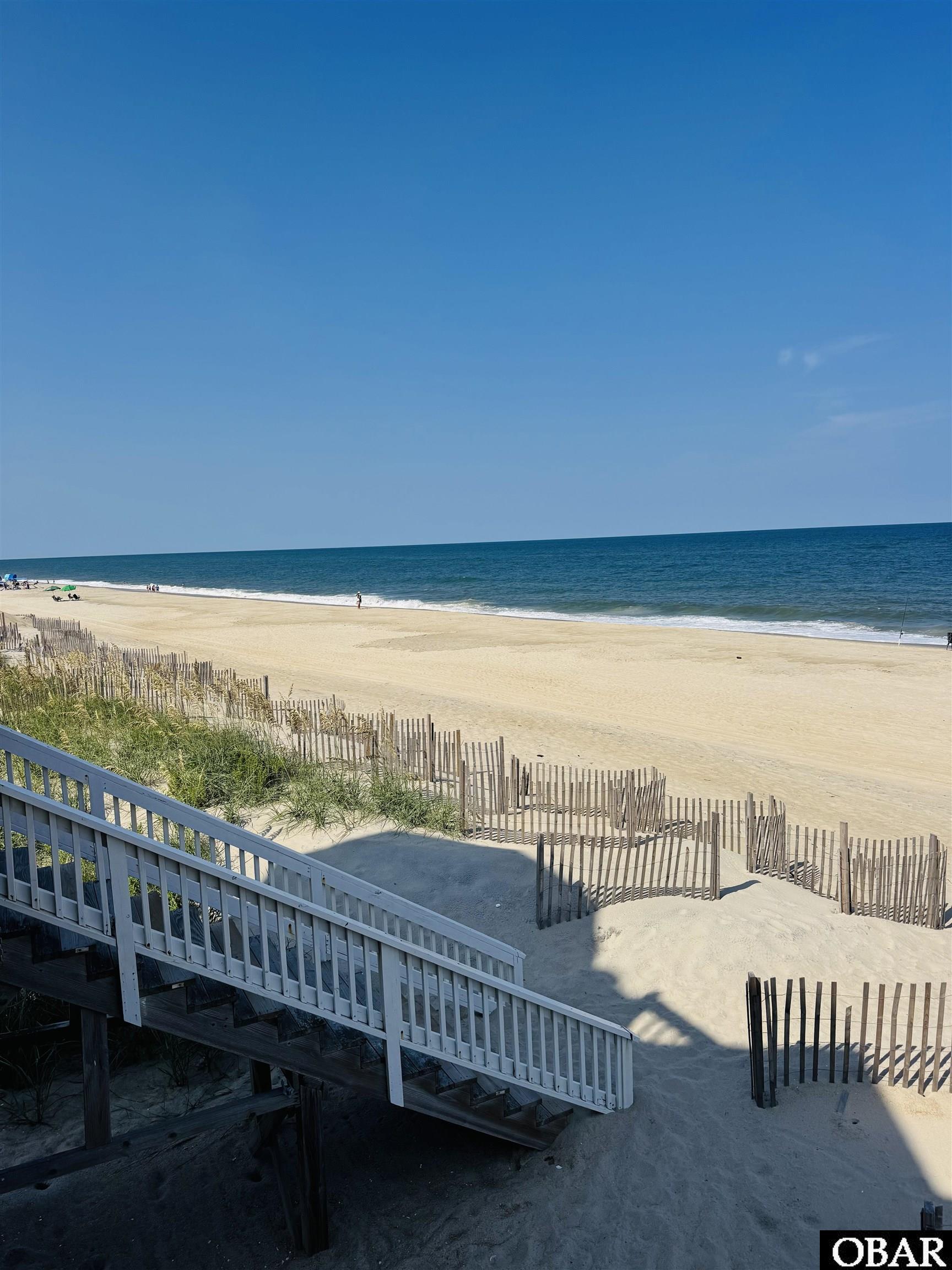 Sandy beach and ocean viewed from a white-railed stairway beside dune grass and sand fencing.