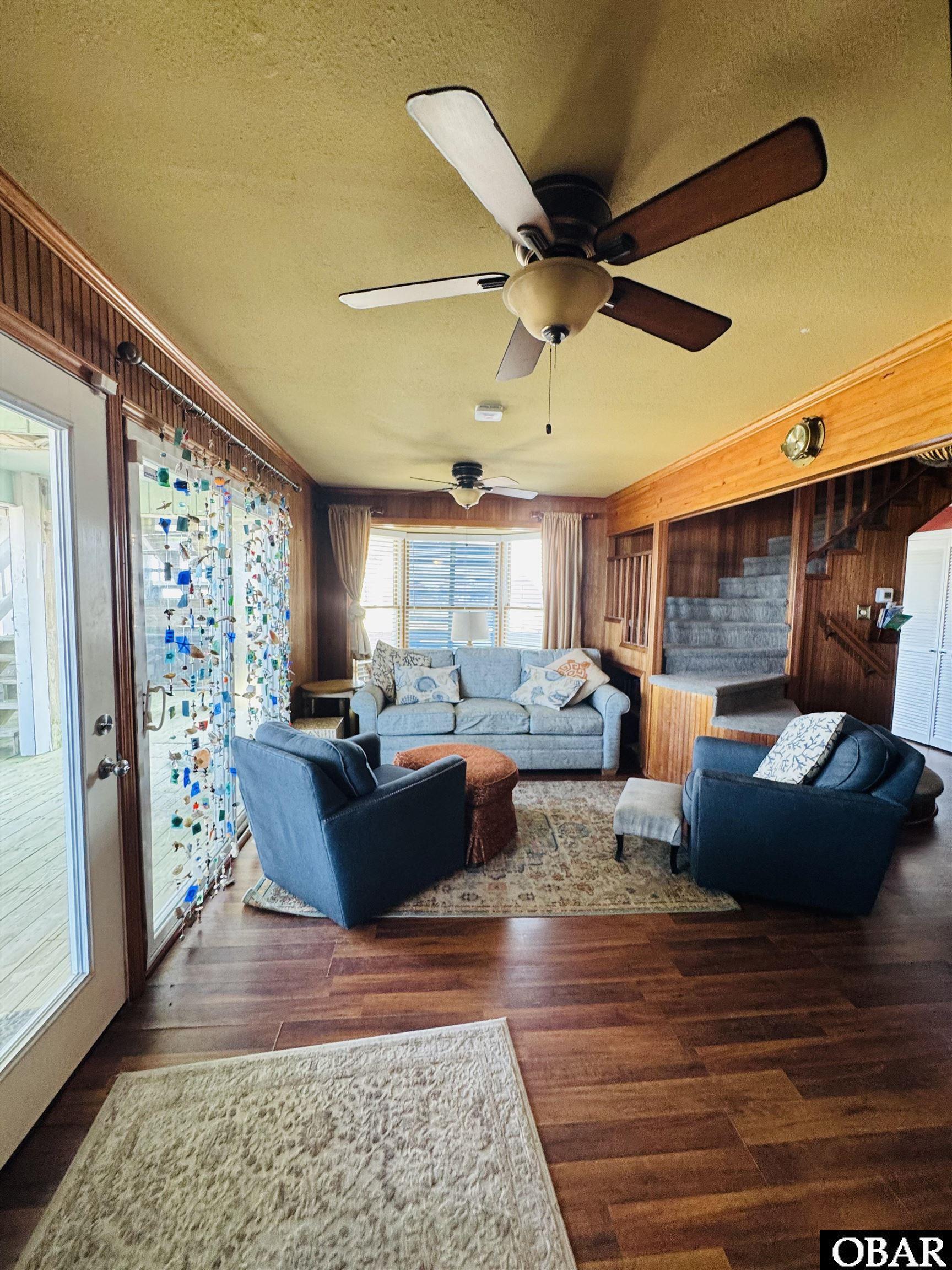 Furnished living room with blue seating, wood-paneled walls, and a carpeted staircase.