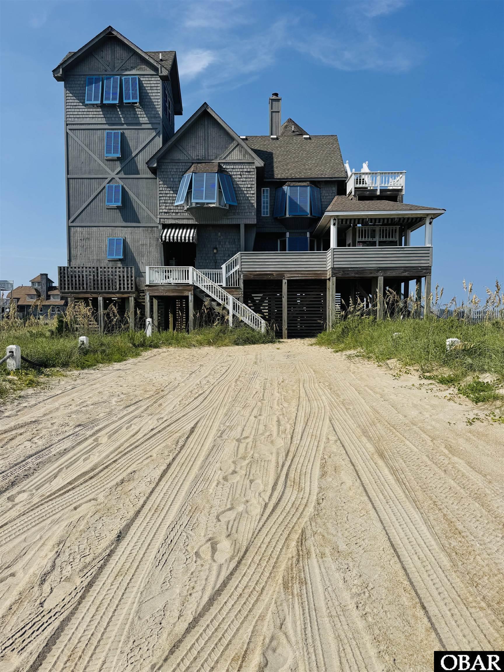 Elevated gray coastal home with blue shutters and multiple decks at the end of a sandy track.