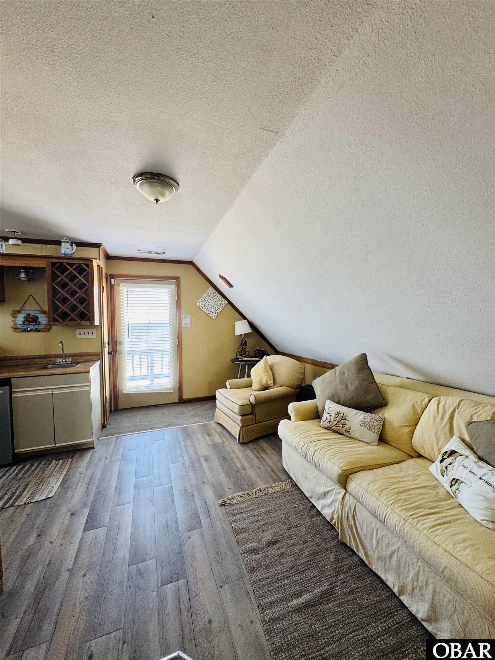 Furnished living room with cream sofas, a compact kitchenette, and a steep sloped ceiling.