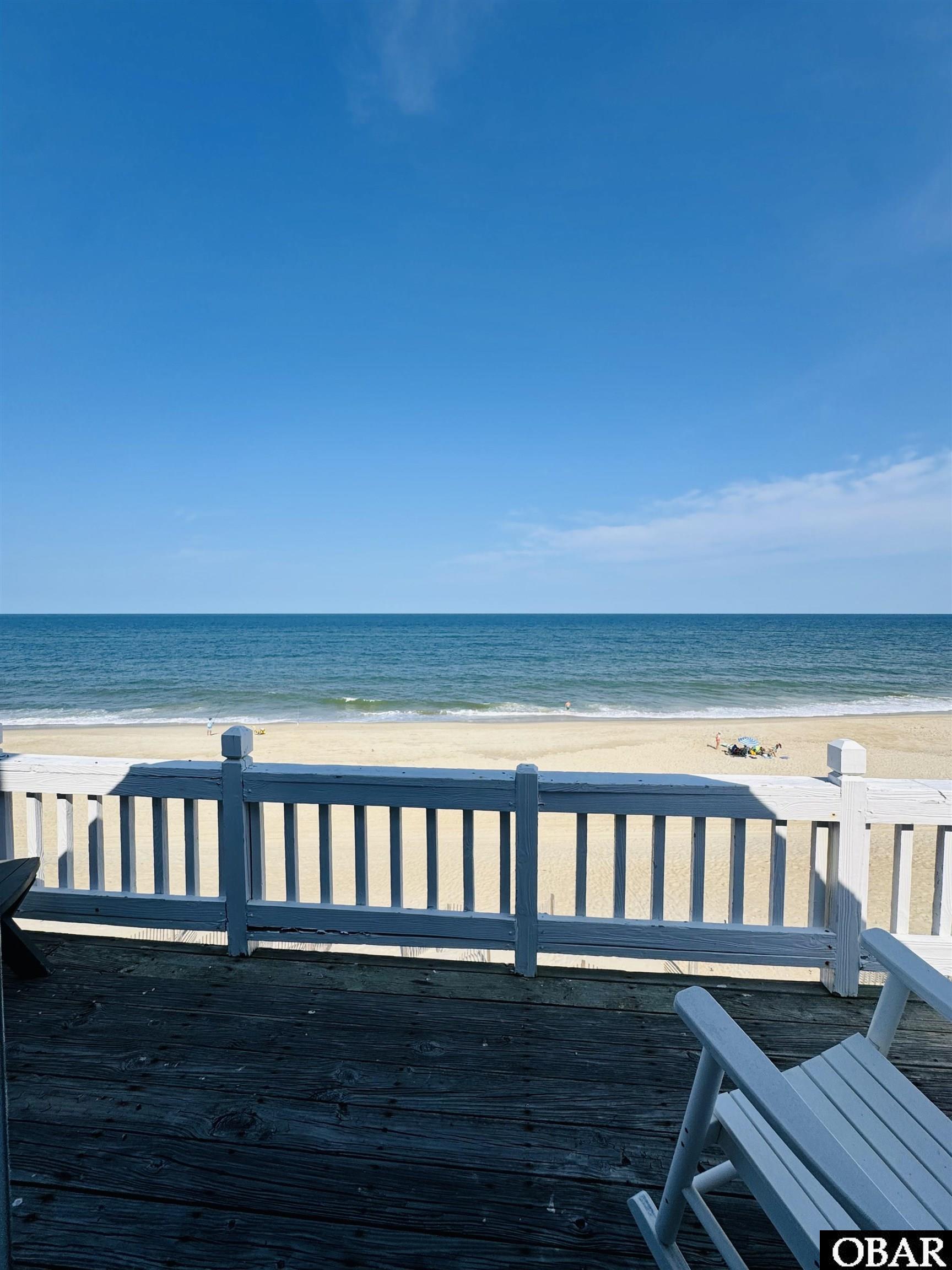 Wooden deck overlooks a sandy beach and ocean, with a white railing and Adirondack chair.