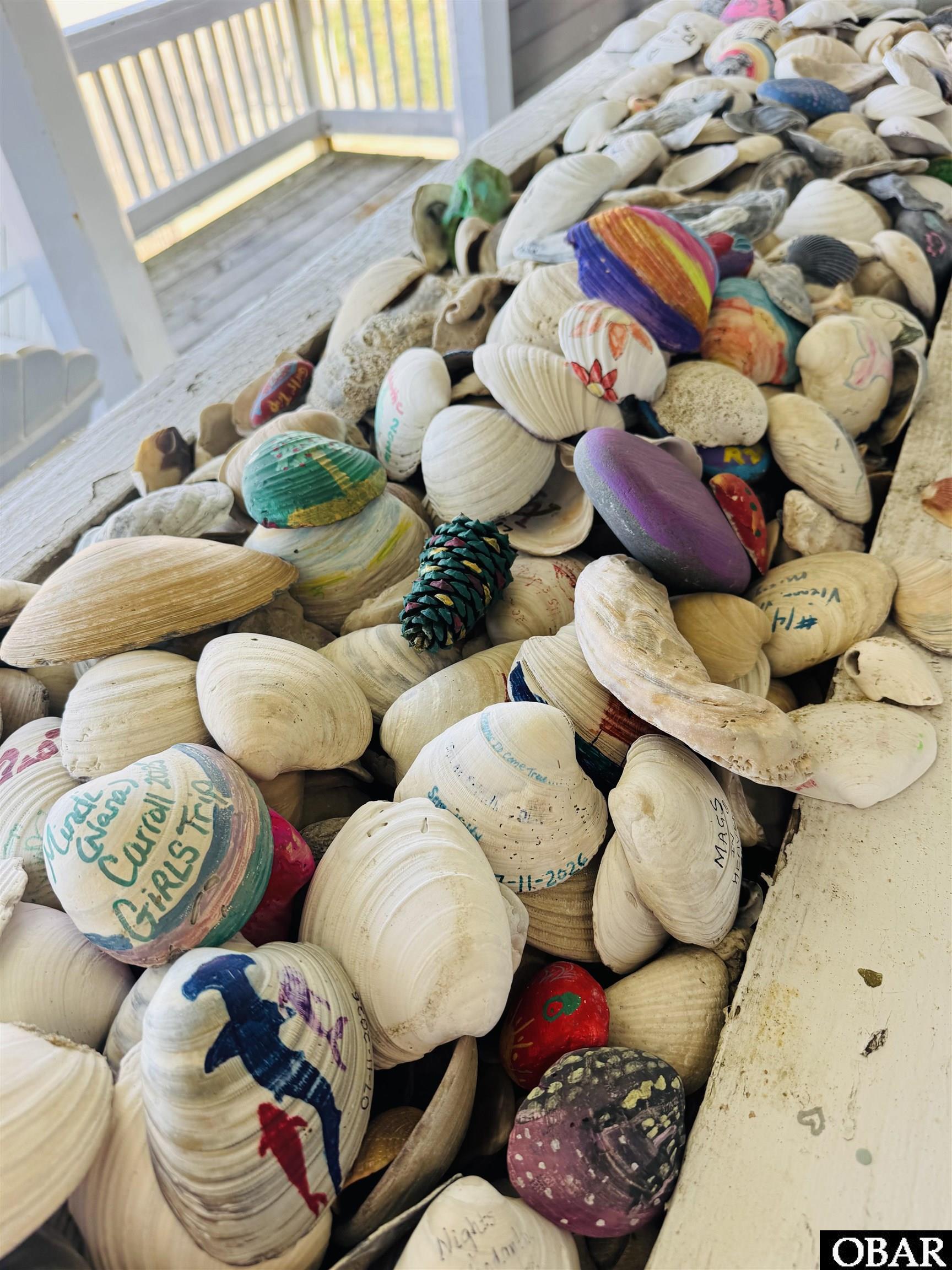 Painted and handwritten seashells piled on a weathered porch beside a white railing.