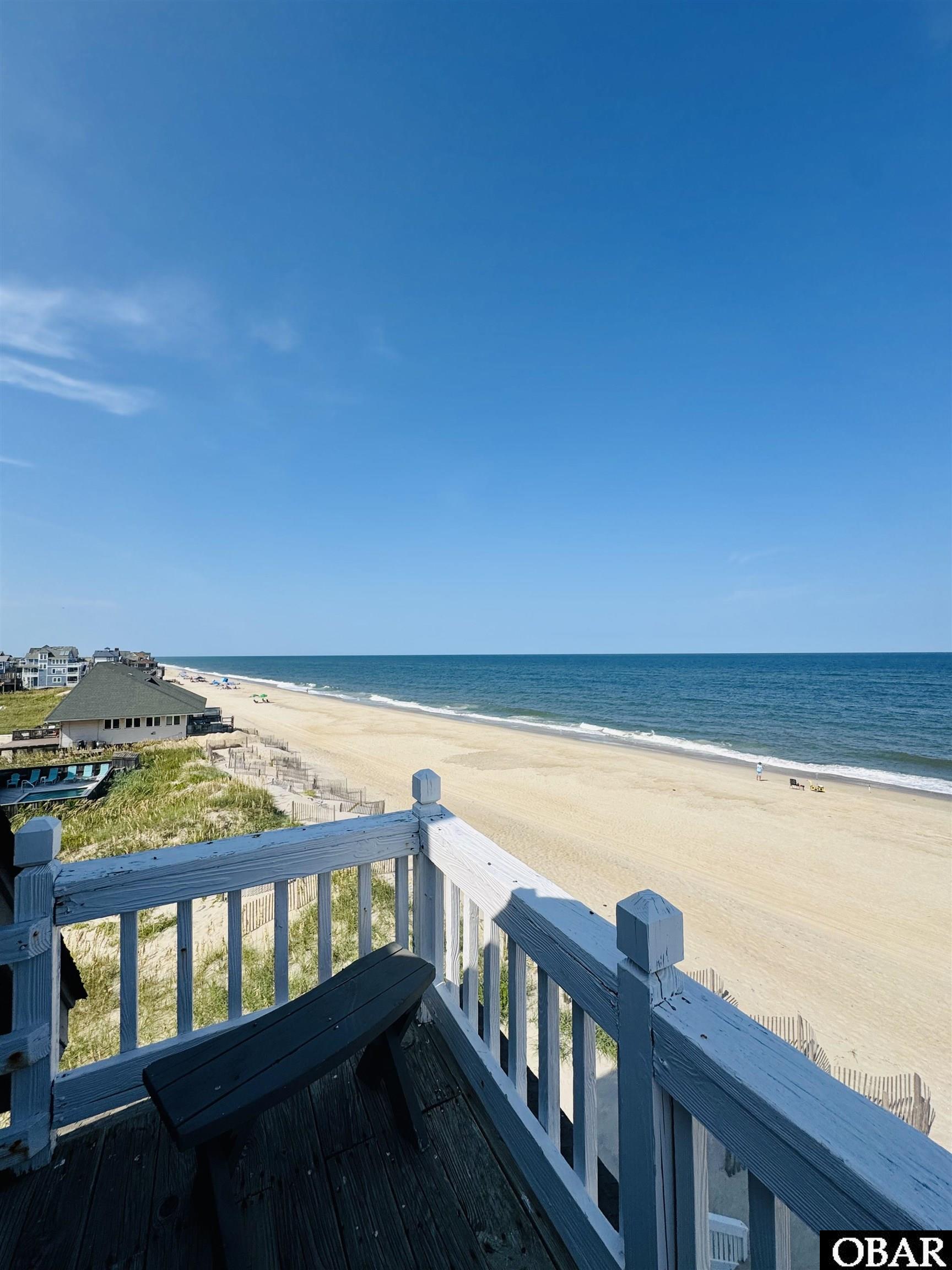 Beach and ocean viewed from a raised deck with white railings and a dark bench.