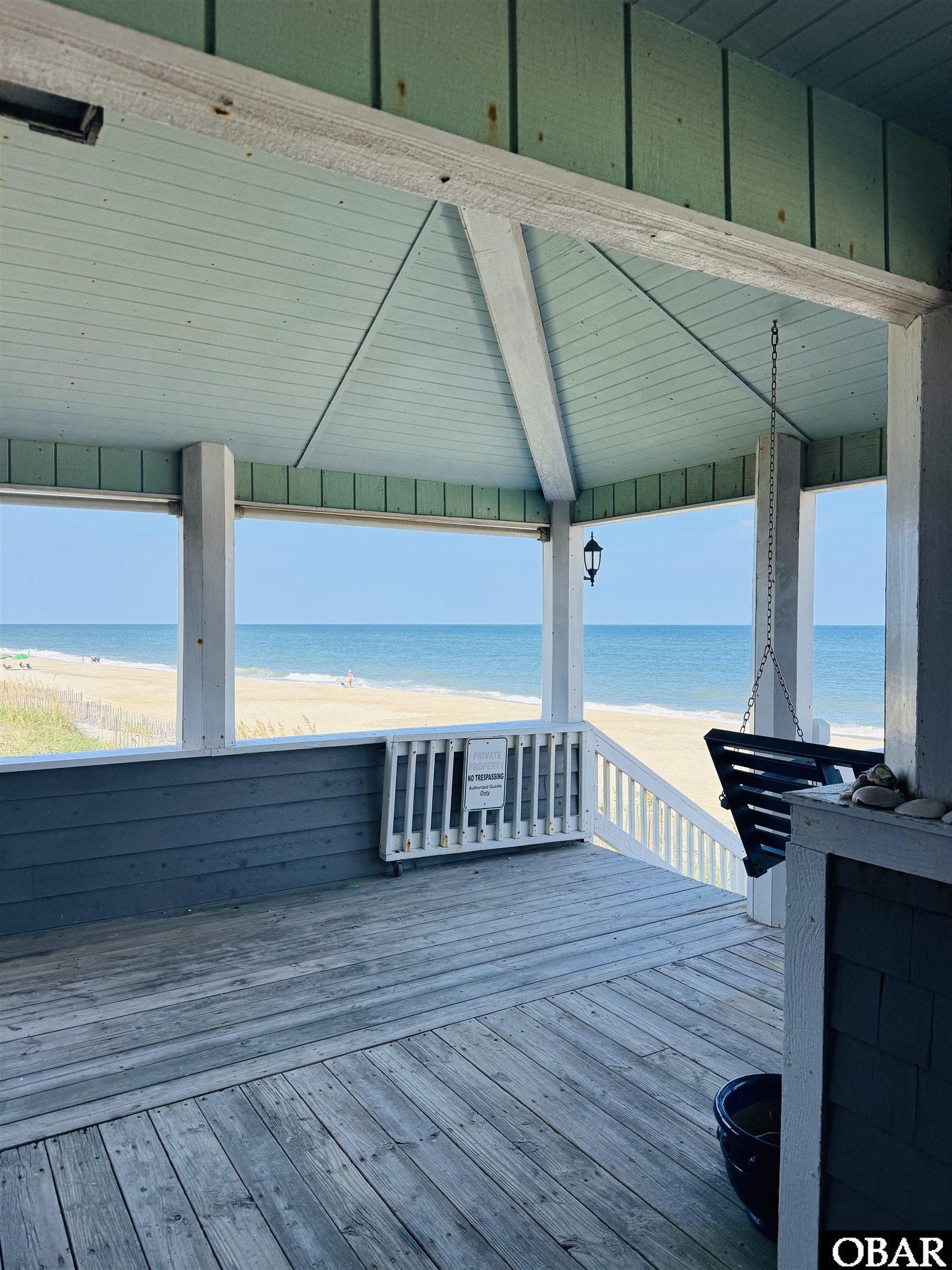 Covered wooden porch with a hanging swing overlooking a sandy beach and ocean.