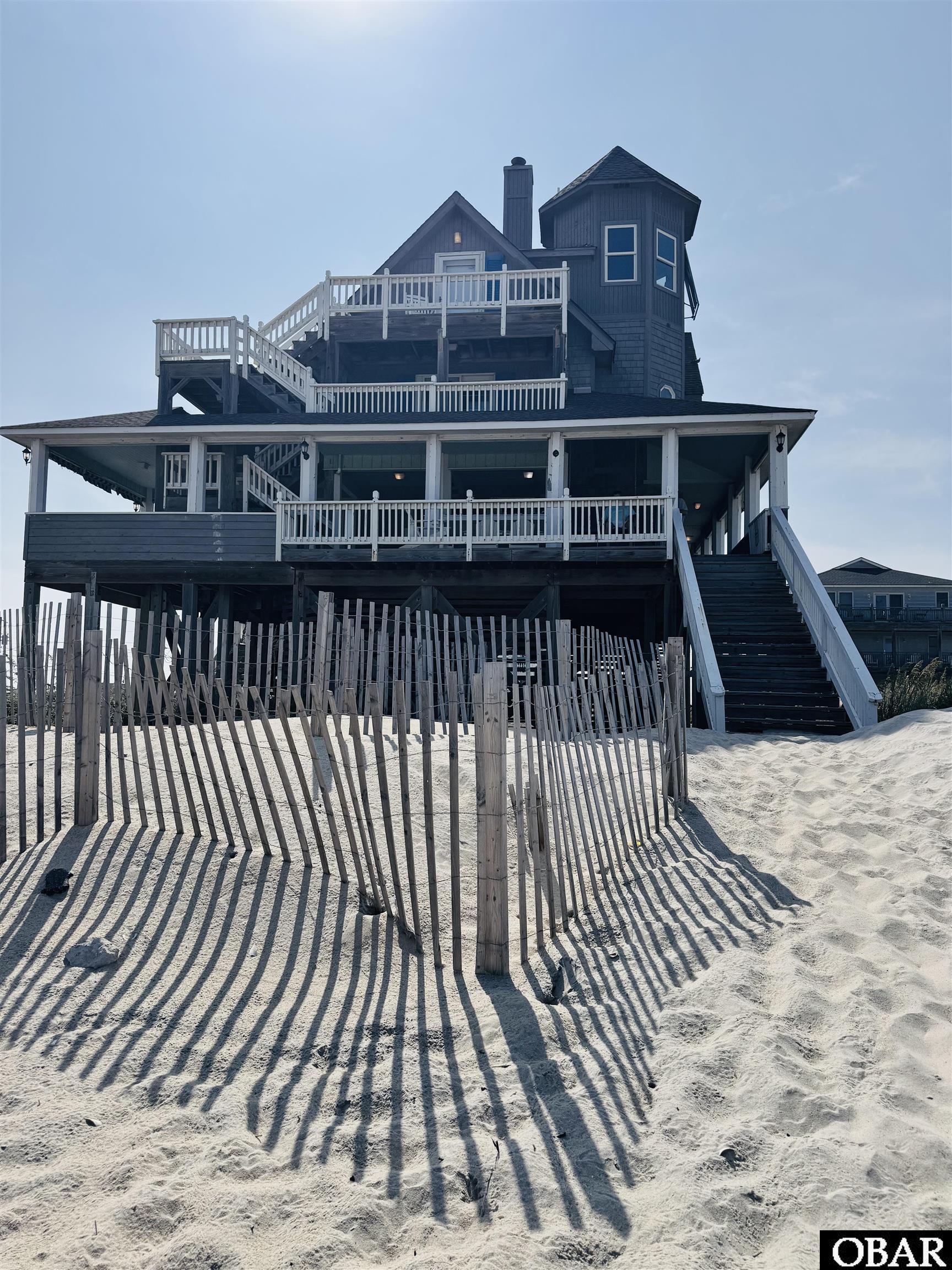 Elevated coastal house with multiple decks and stairs behind a dune fence on sandy ground.