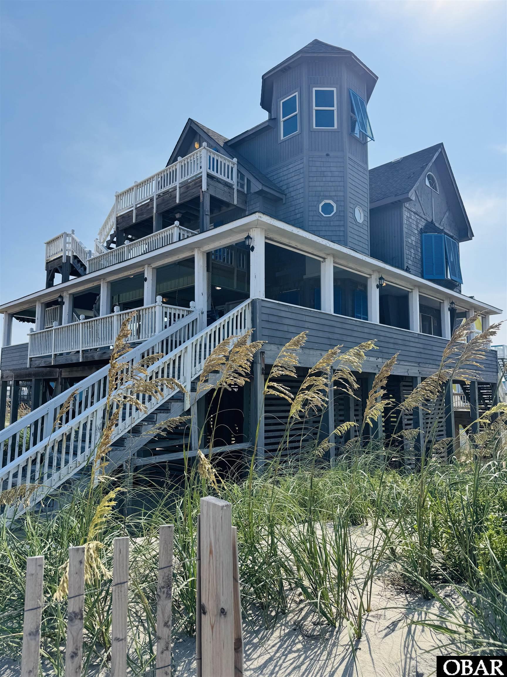Elevated coastal house with white decks and stairs rises behind beach grass and a sandy path.