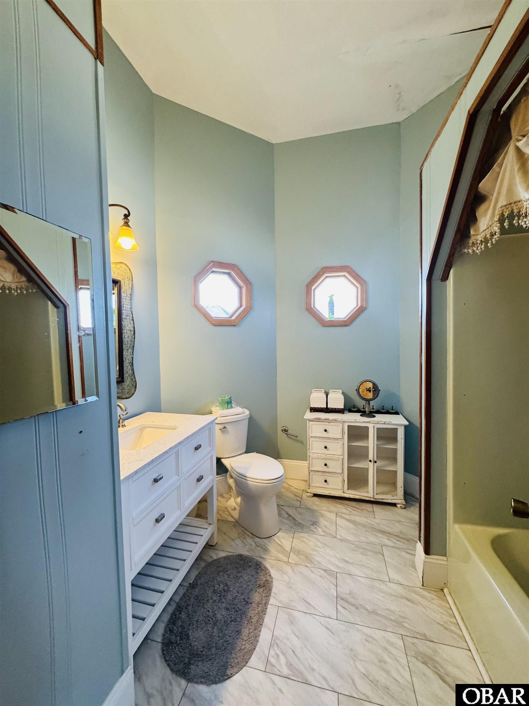 Bathroom with pale blue walls, two octagonal windows, a white vanity, and marble-look tile flooring.