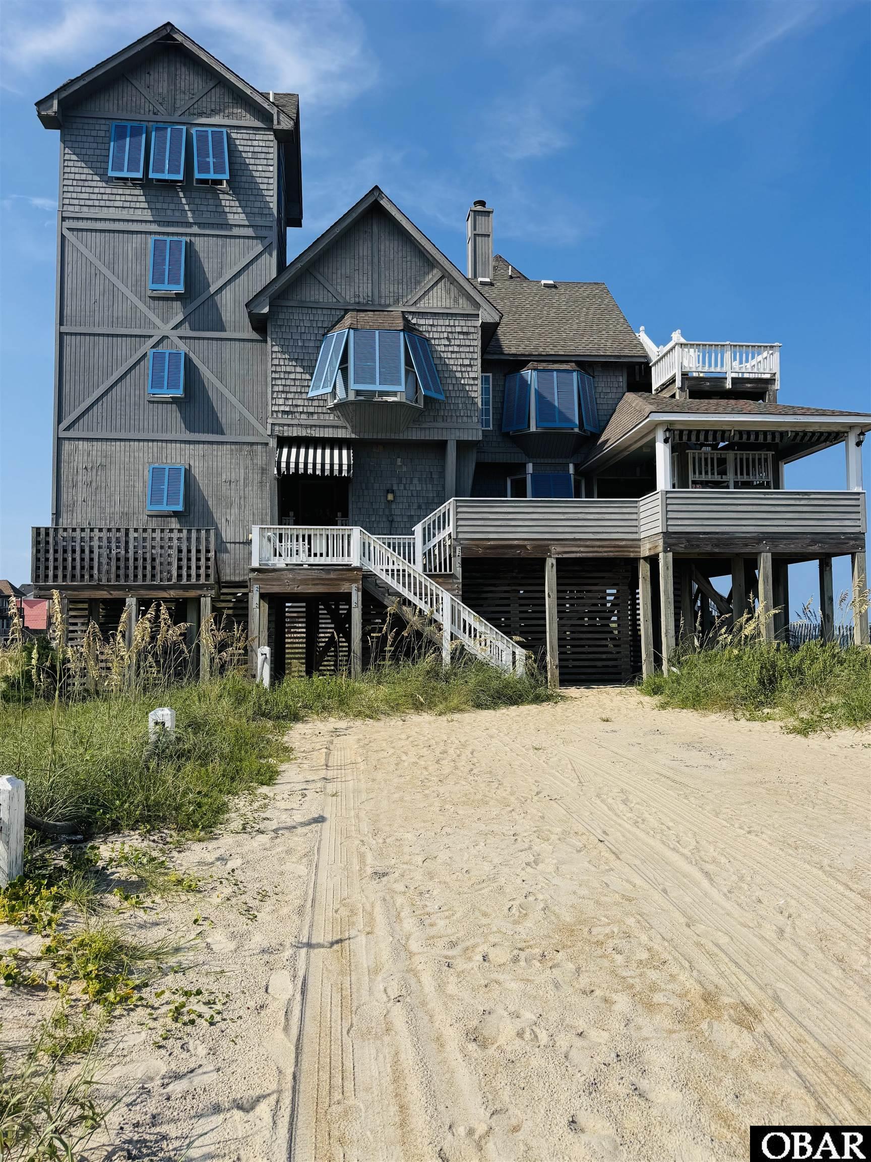 Elevated gray coastal home with blue shutters, wraparound decks, and a sandy approach.