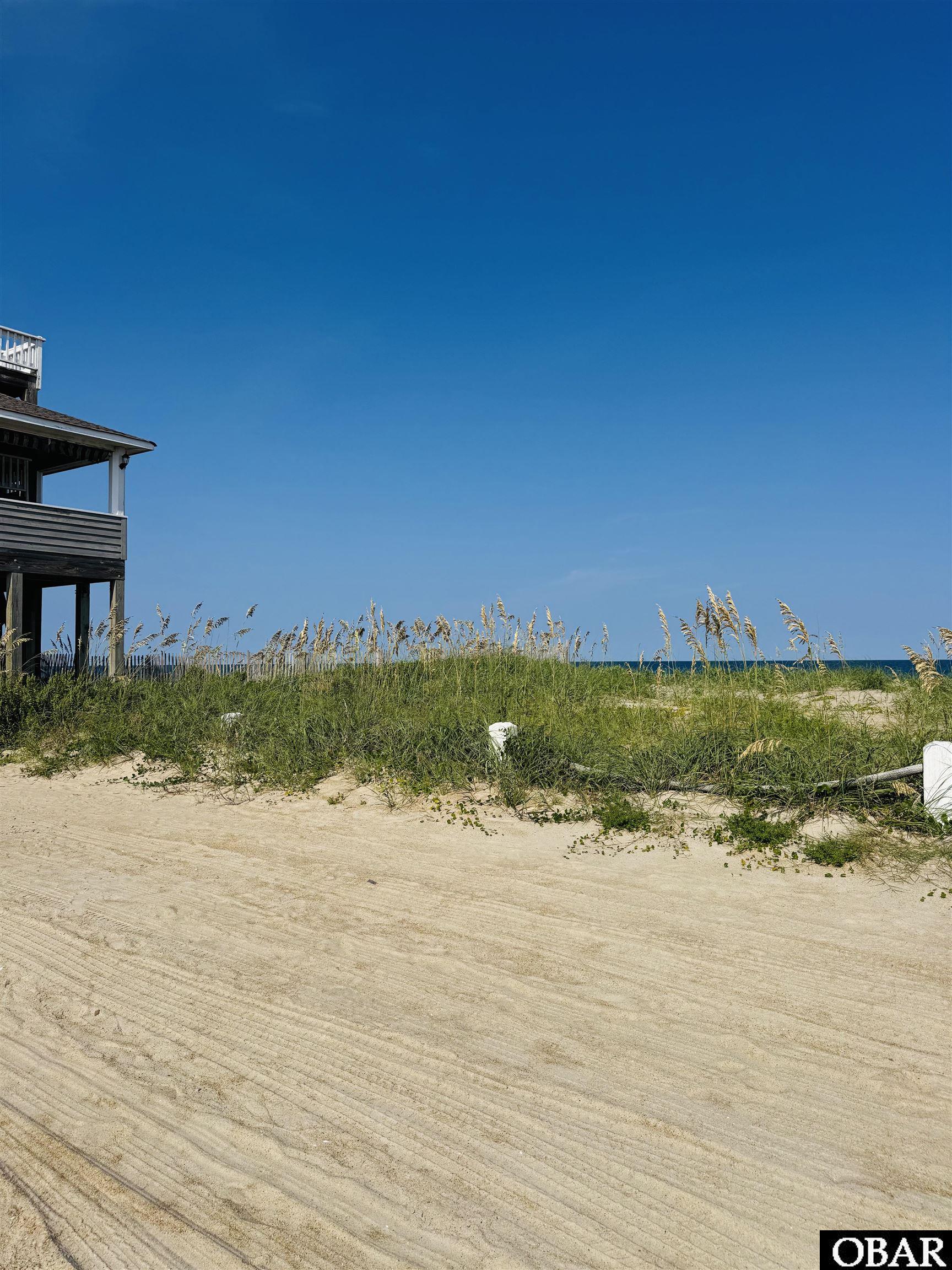 Beachfront lot with dune grass, ocean, sandy access road, and part of an elevated house.