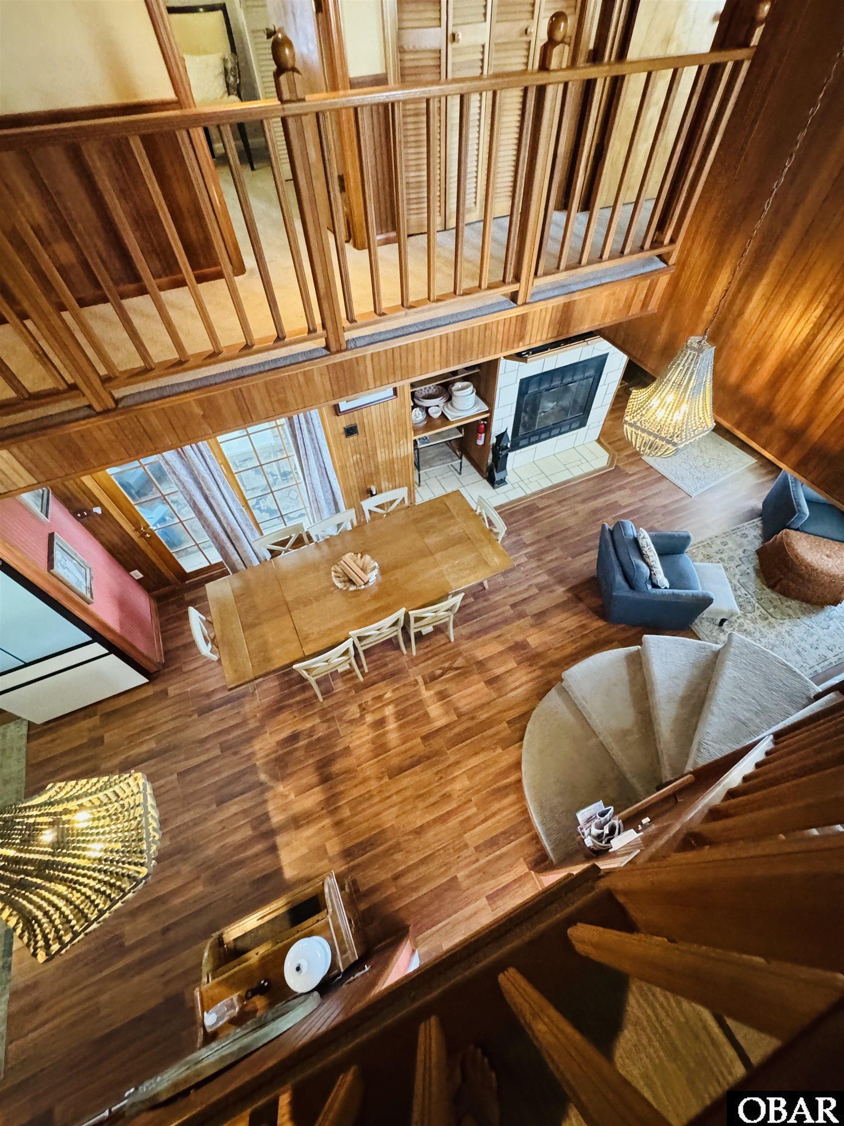 Open wood-paneled living and dining room viewed from above, with a tiled fireplace and curved staircase.