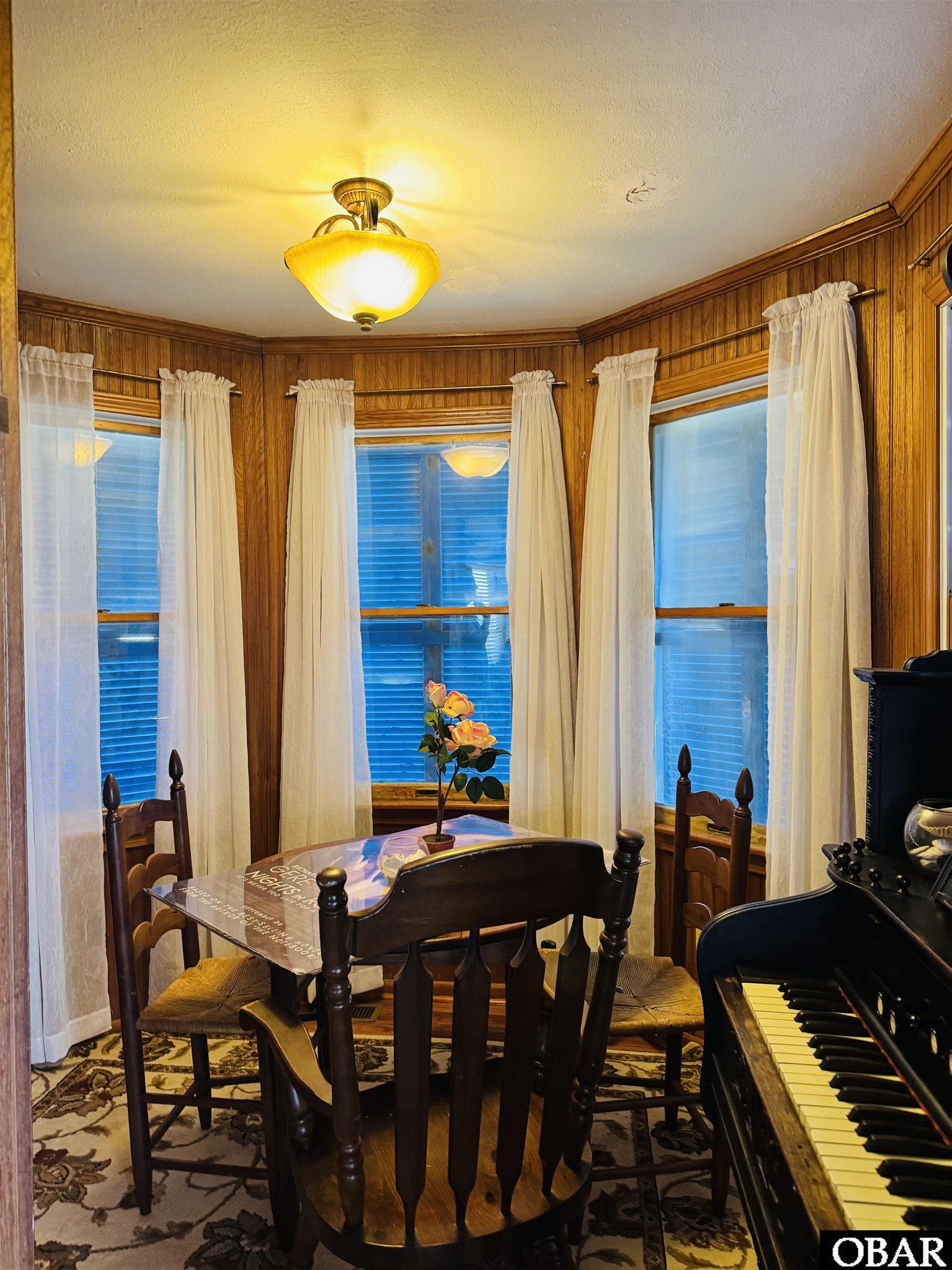 Dining room with wood-paneled walls, a round table, curtained windows, and a black piano.