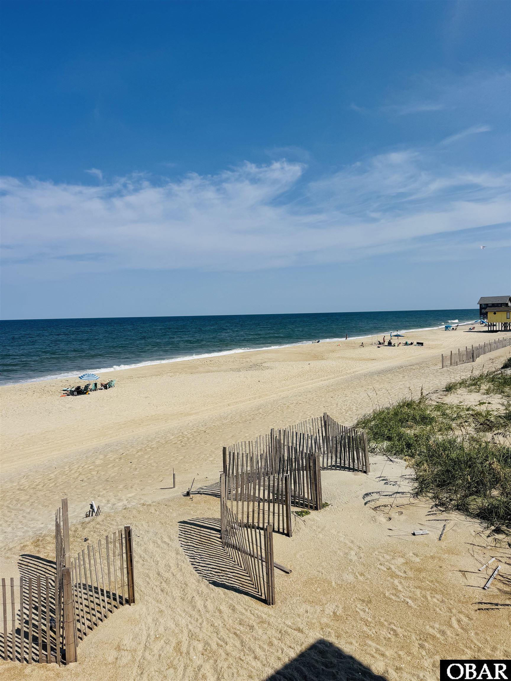 Sandy ocean beach with wooden sand fences in the foreground and an elevated building along the shore.