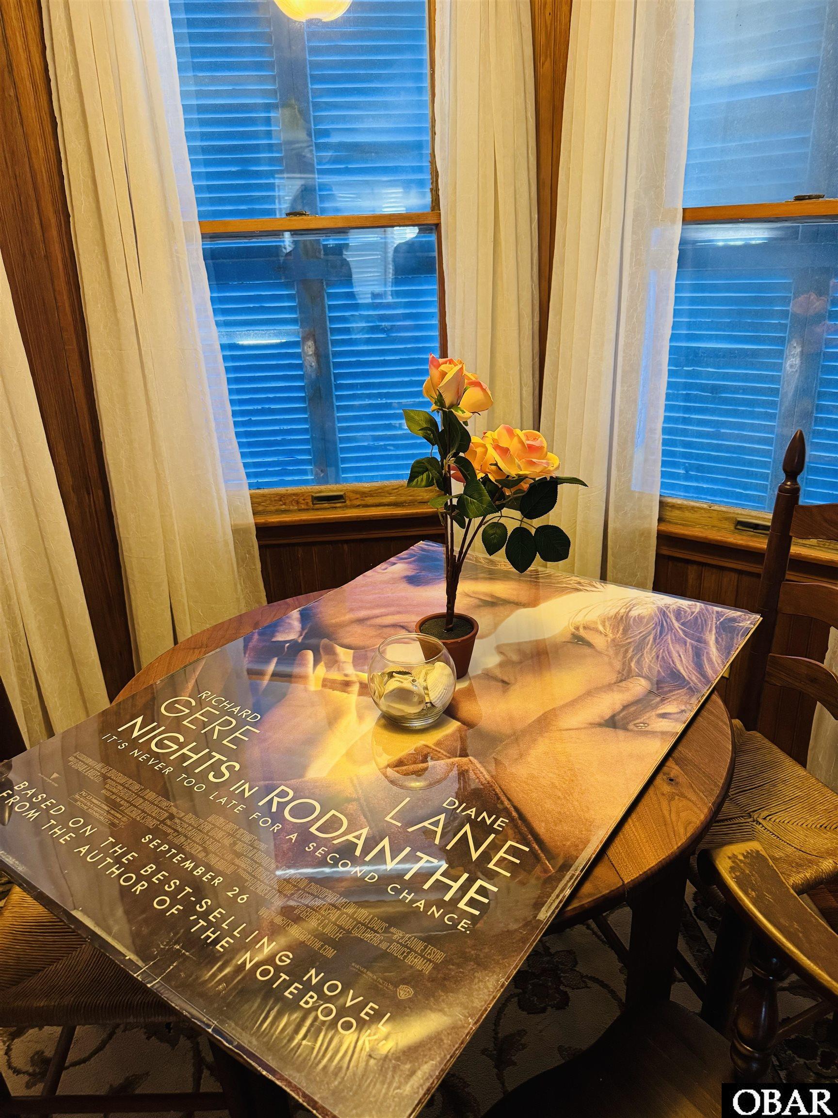 Dining nook with a movie poster covering the table, a potted rose, and blue-shuttered windows.