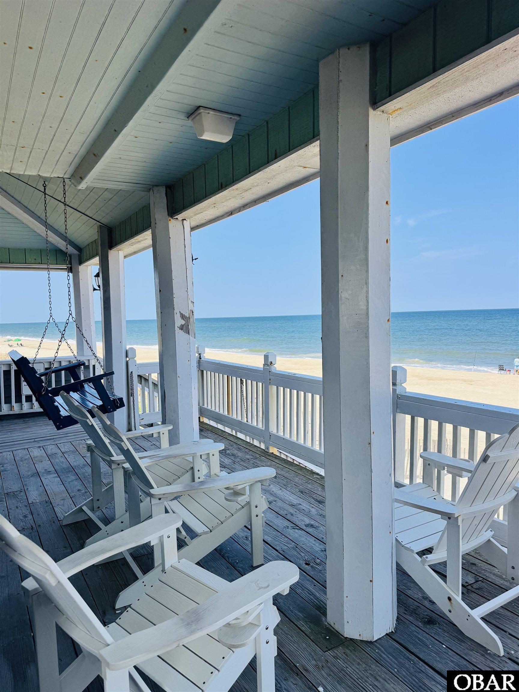 Covered oceanfront porch with white Adirondack chairs and a hanging swing overlooking the beach.