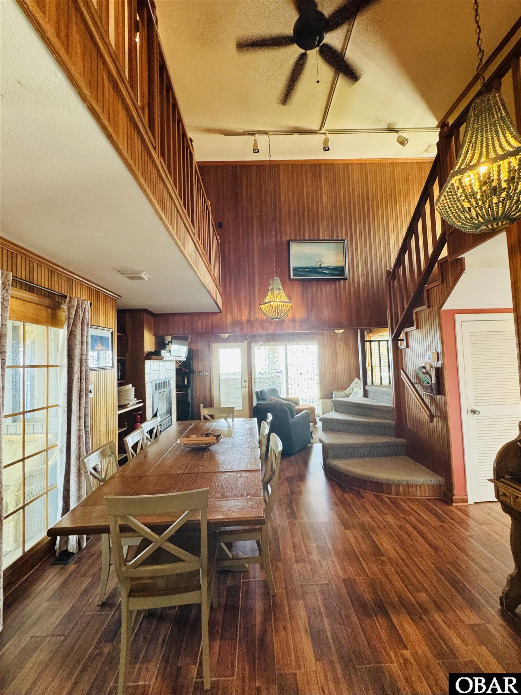 Open-plan dining and living room with wood paneling, a long table, fireplace, and carpeted staircase.