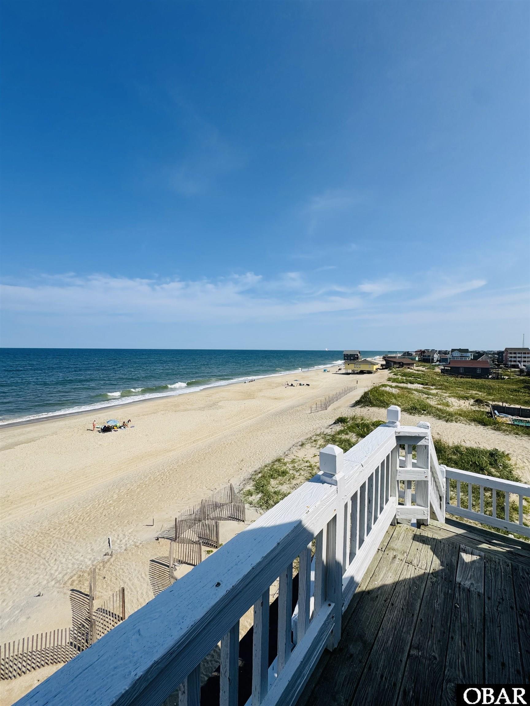 Oceanfront beach view from a wooden deck, with white railings and houses along the shore.