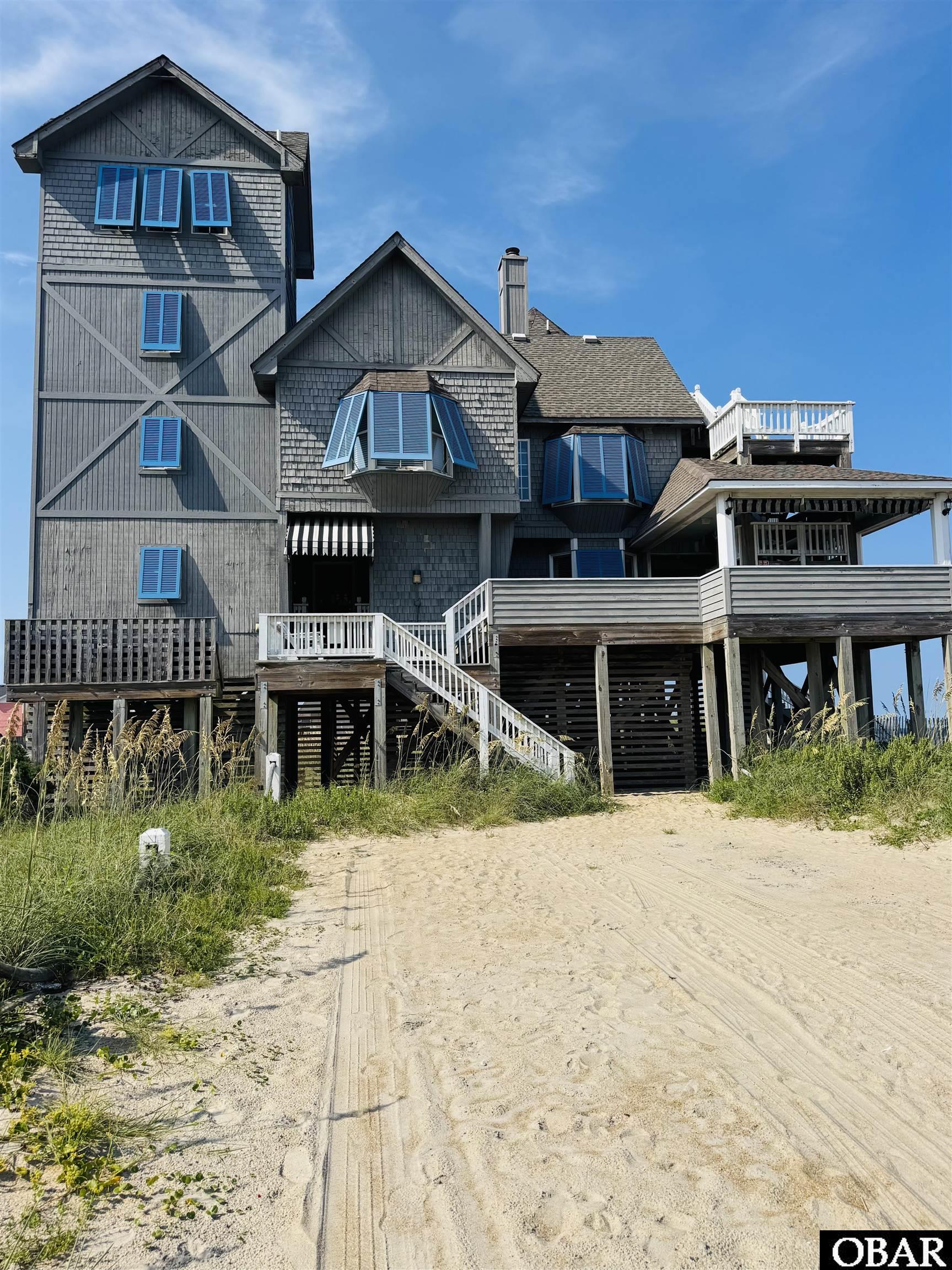 Elevated gray beach house with blue shutters, white-railed decks, and a sandy approach.