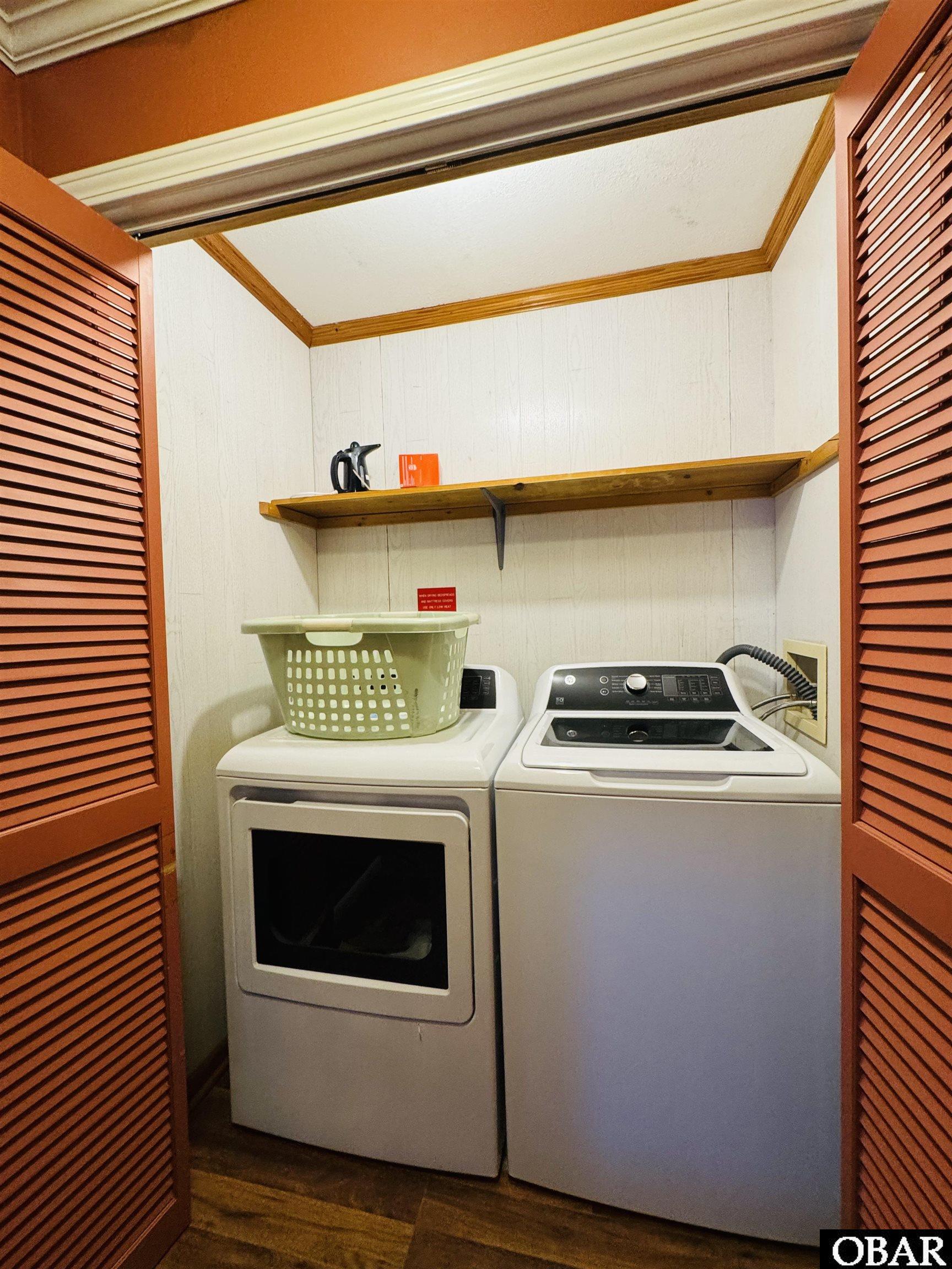 Laundry closet with a dryer, top-loading washer, green basket, and wooden shelf.