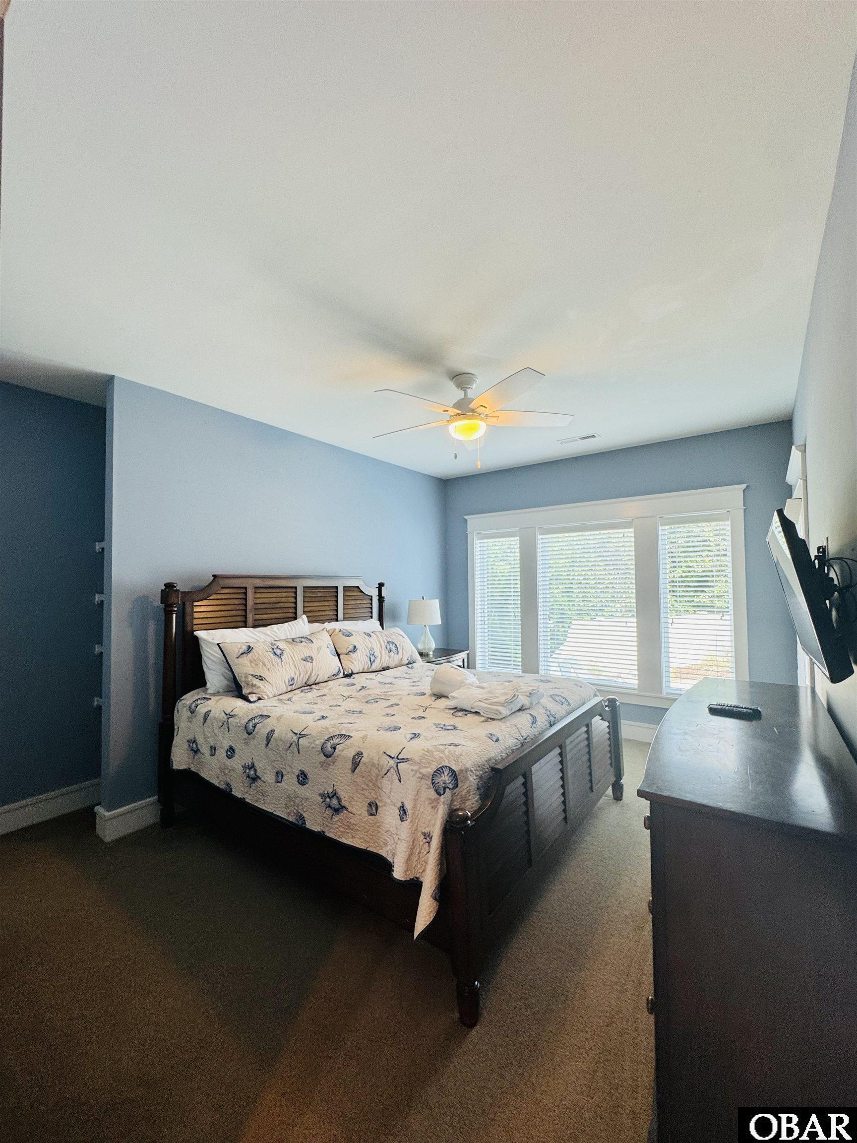 Bedroom with blue walls, seashell-patterned bedding, a ceiling fan, and wide windows with blinds.