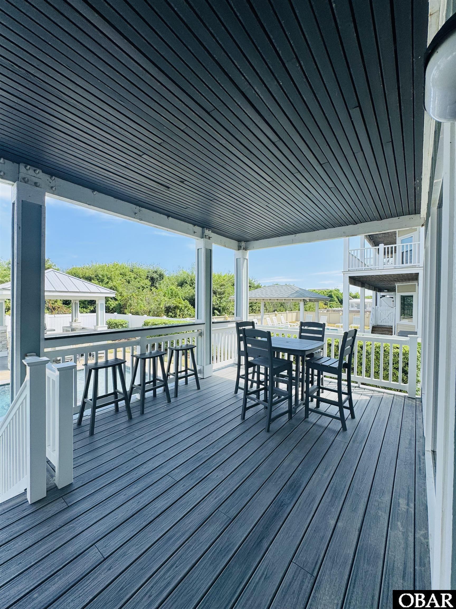 Covered deck with black dining set and bar stools overlooking a pool and gazebo.