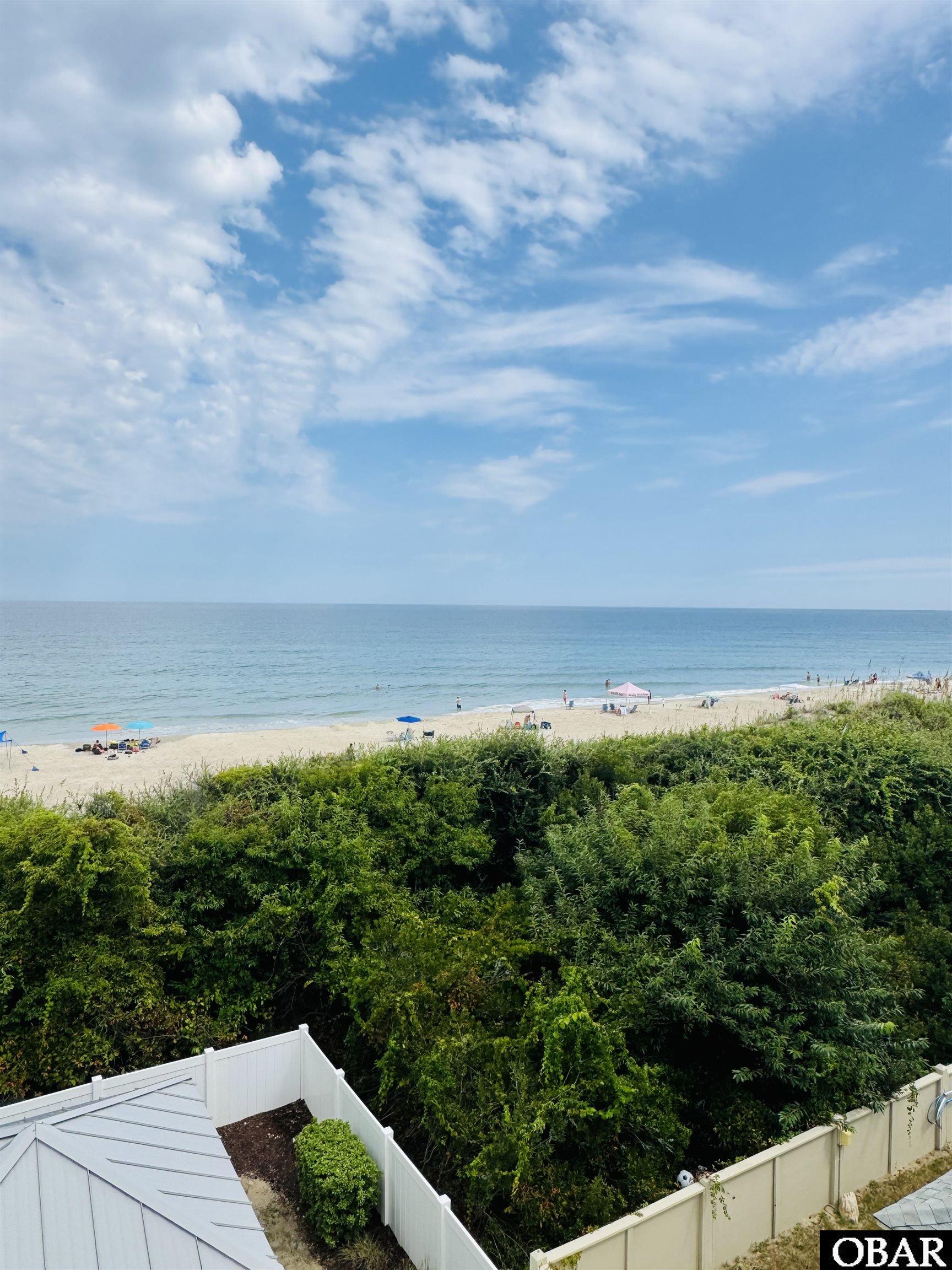 Ocean and sandy beach beyond dense shrubs, with a white fence and roof in the foreground.