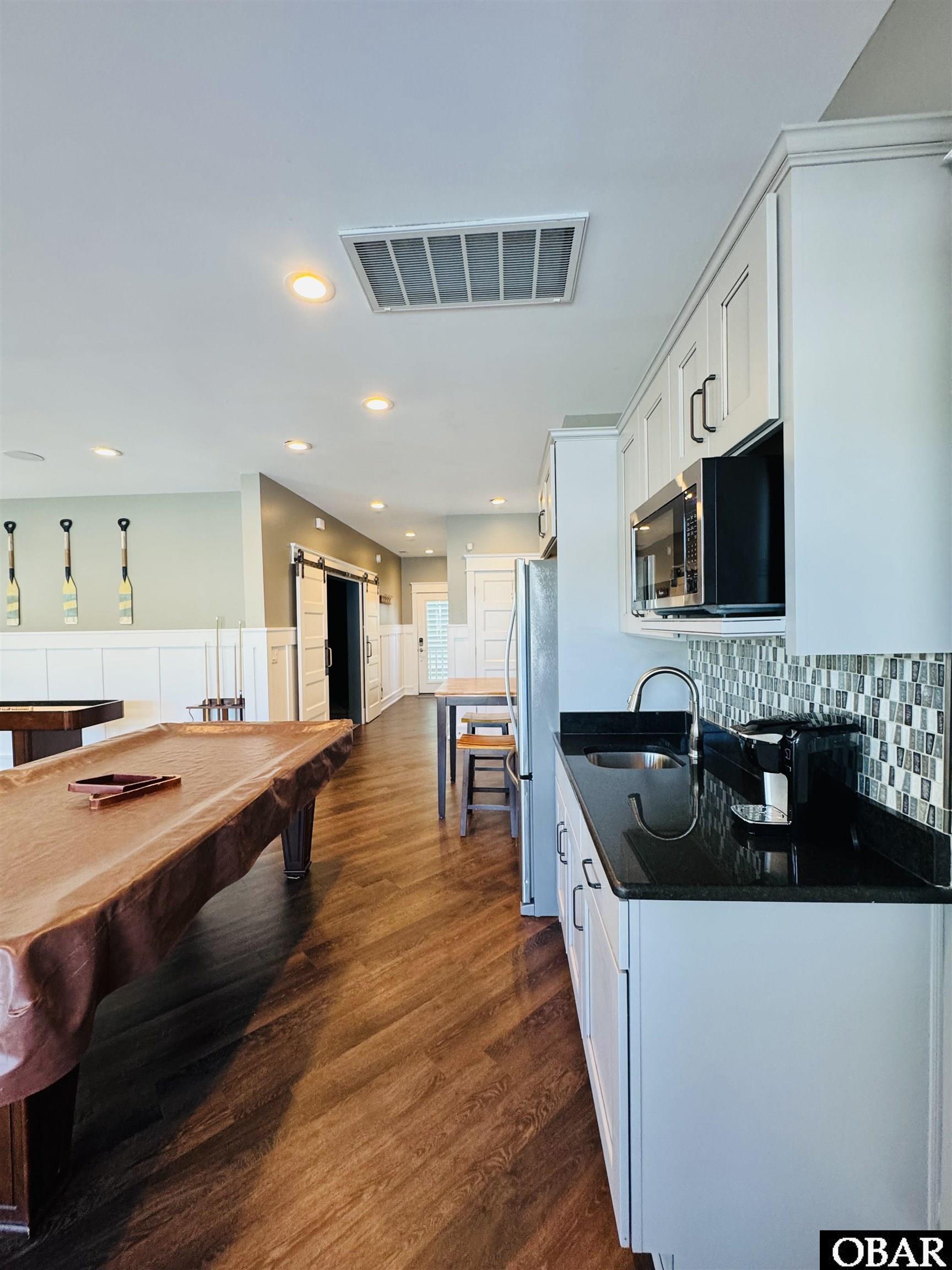 Kitchen with white cabinets and black counters, opening to a room with a large pool table.
