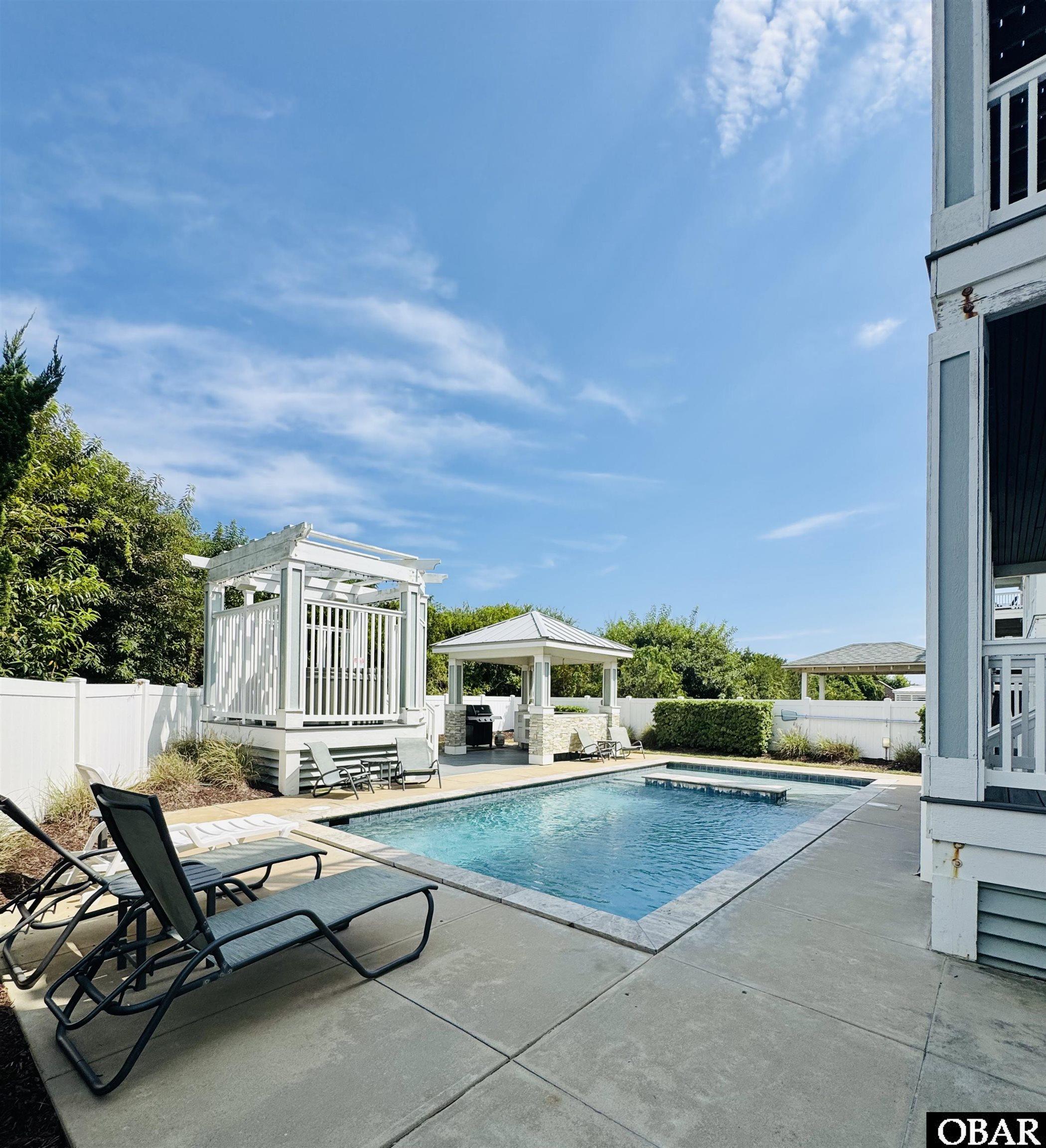 Outdoor pool with lounge chairs, a raised white pergola, and a covered grilling pavilion.