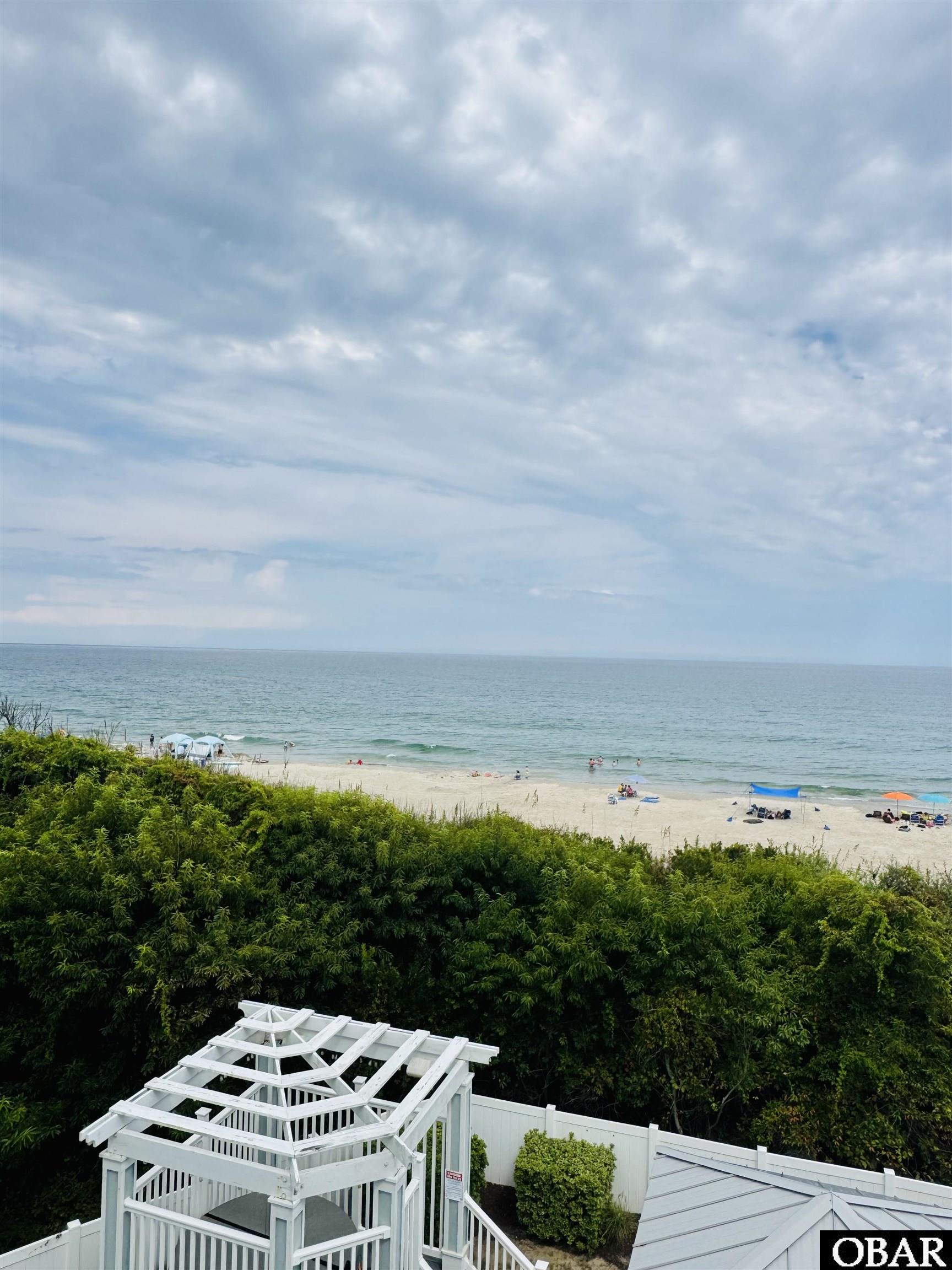 Oceanfront beach with swimmers and umbrellas, viewed over greenery and a white pergola.