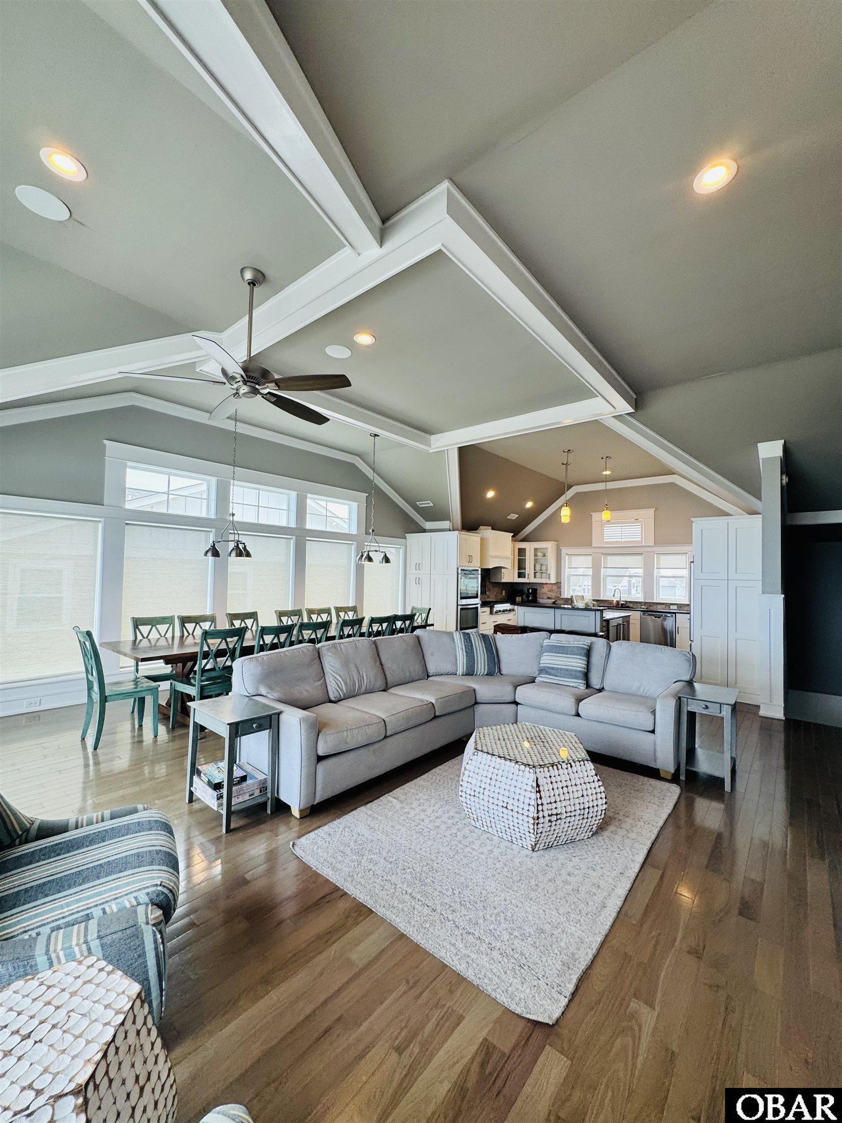 Open-plan living room with a gray sectional, hardwood floors, and vaulted ceiling beside the dining area and kitchen.