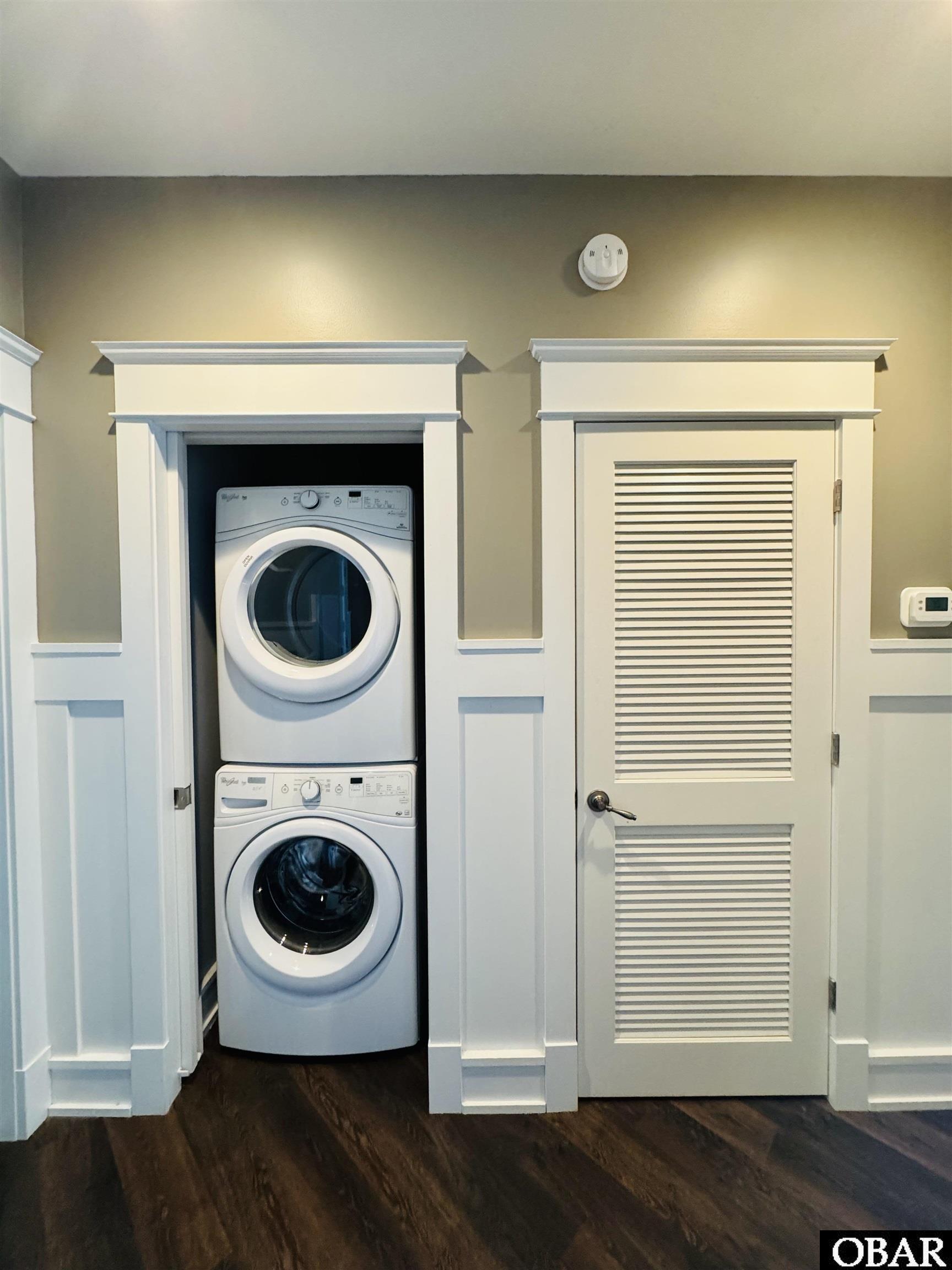 Stacked white washer and dryer beside a built-in louvered door in a trimmed laundry area.