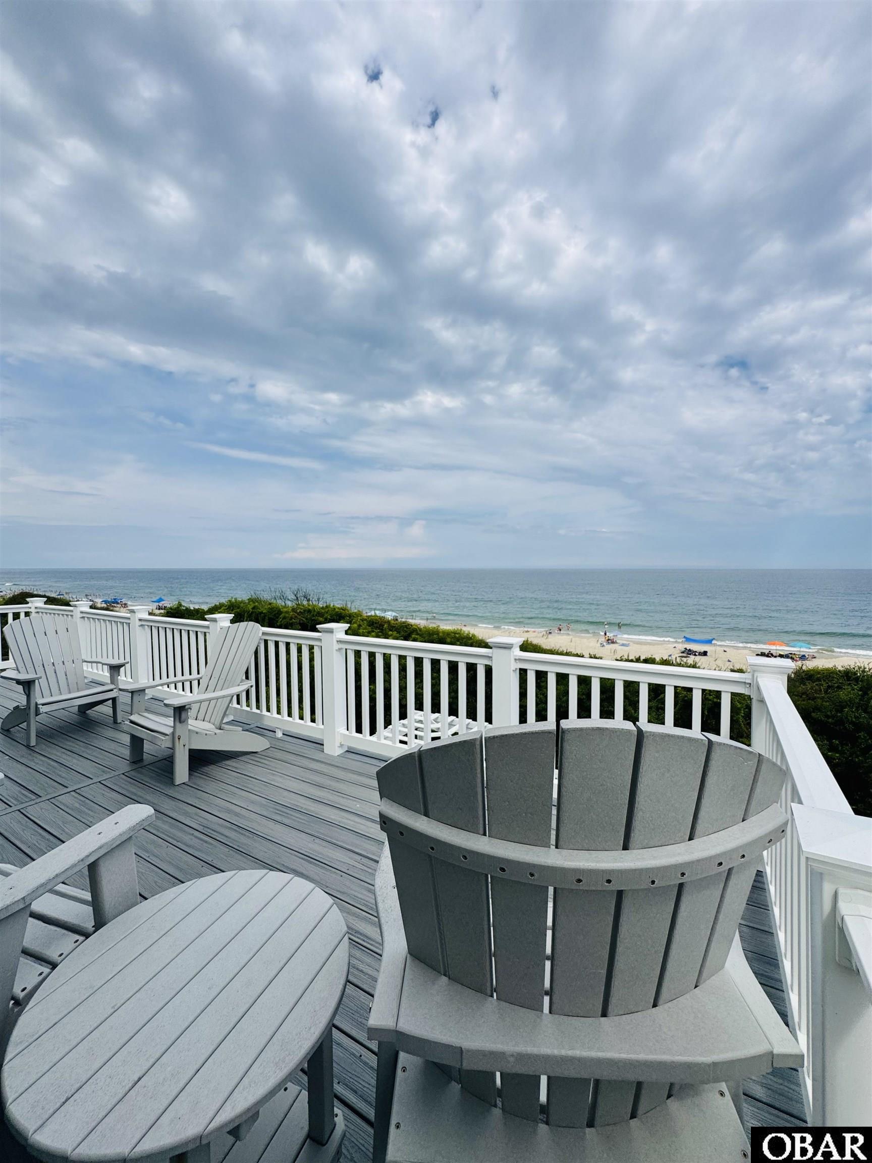 Ocean-view deck with gray Adirondack chairs, white railing, and beach beyond.