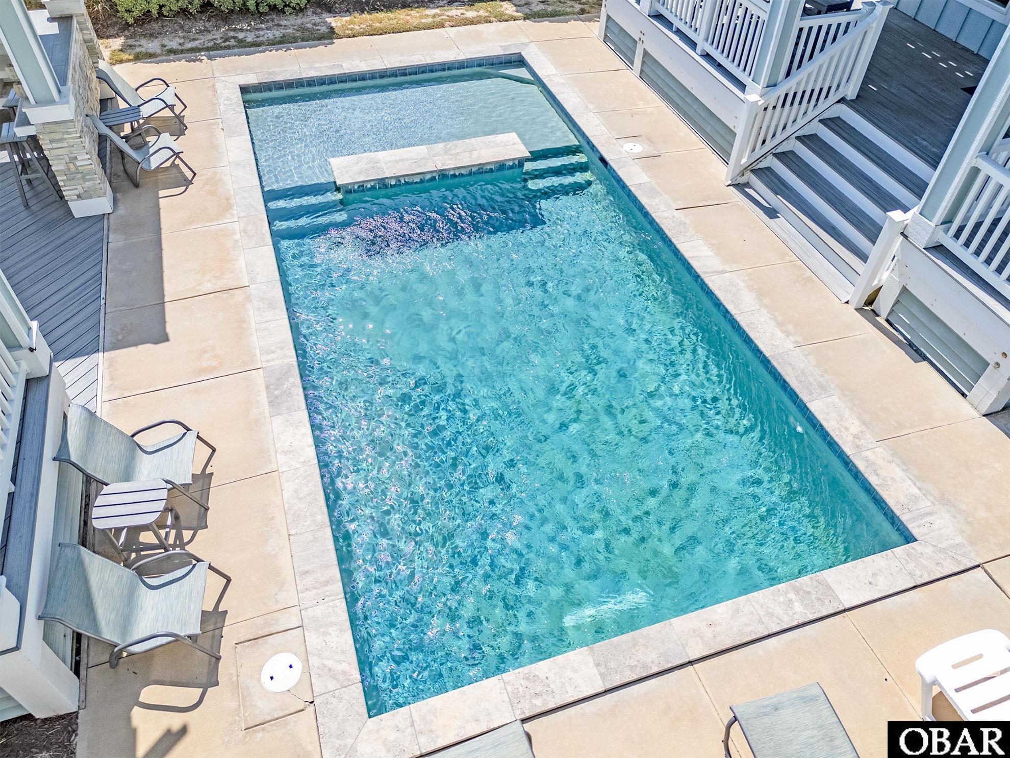Outdoor pool with a shallow ledge and raised spillover feature, surrounded by a stone deck and patio chairs.