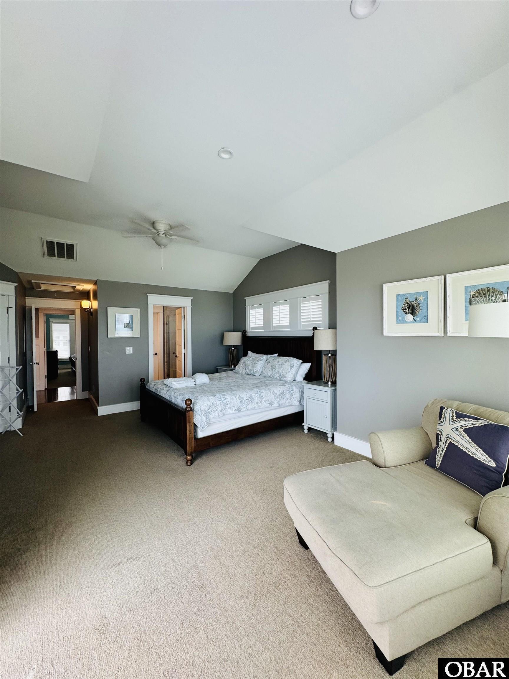 Bedroom with a king bed beneath a vaulted ceiling and a cream chaise lounge.