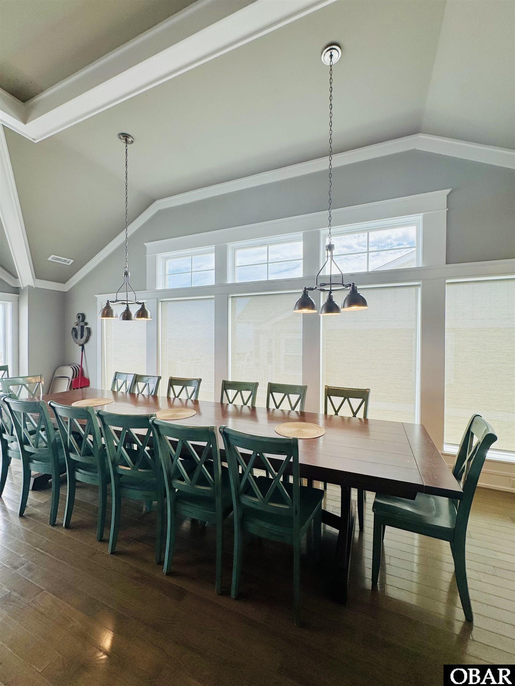 Dining room with a long table, green chairs, vaulted ceiling, and large windows.