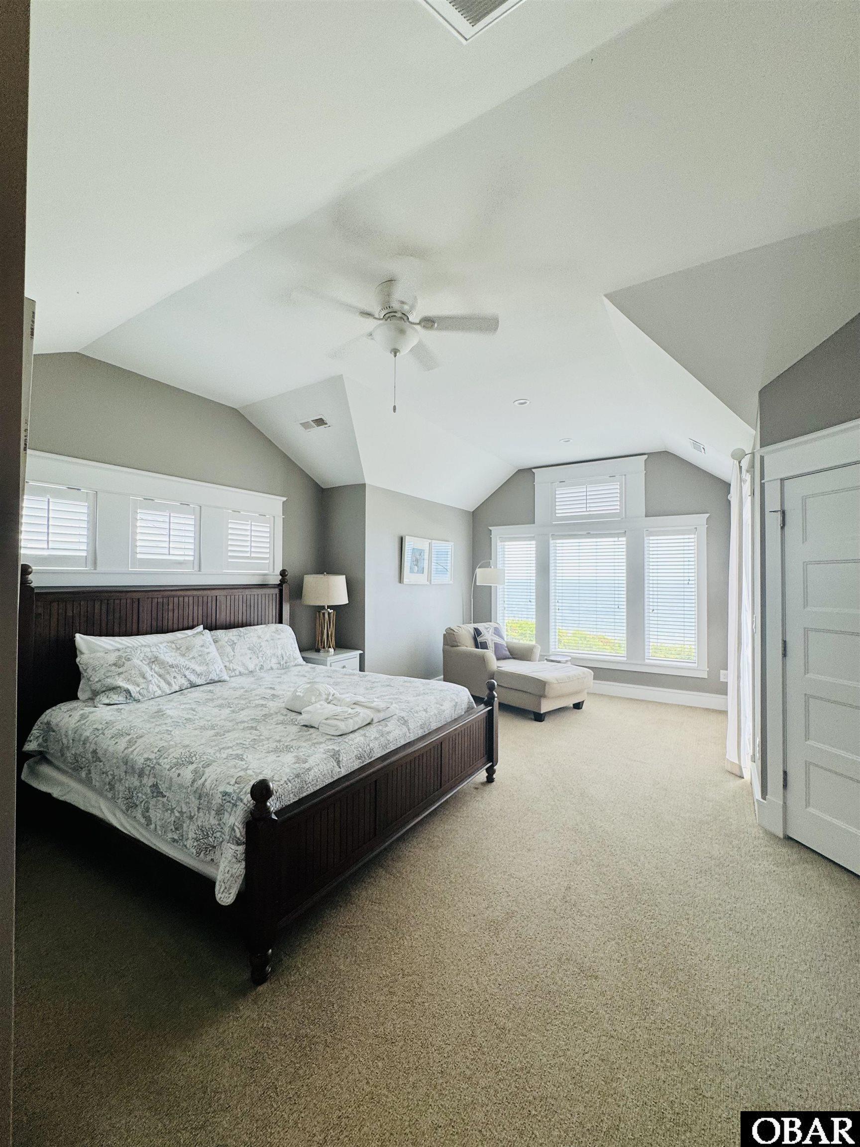Furnished bedroom with a dark wood king bed, vaulted ceiling, and chaise beside shuttered windows.